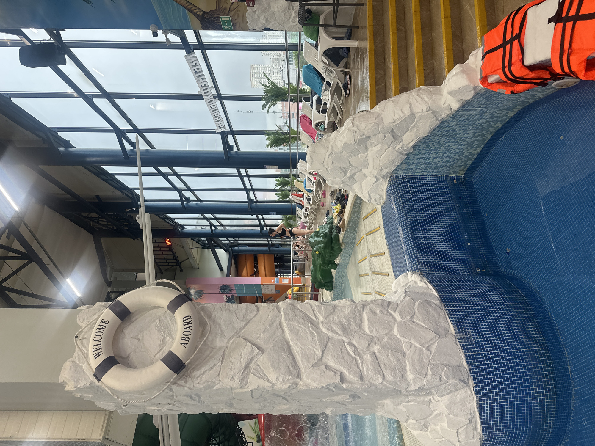 This image depicts an indoor water park facility, likely located in Moscow, Russia, given the context of the surrounding city visible through the large windows. The foreground features a section of a pool with blue tiled walls and bottom. A white, rock-like structure, decorated with a life preserver that reads "WELCOME ABOARD," stands to the left. To the right, there are steps leading up to a tiled platform where several orange life vests are placed.
In the mid-ground, a person in a black swimsuit is standing, facing away from the camera. Further back, a row of white lounge chairs is arranged, some with colorful towels draped over them, suggesting a relaxation area. The background is dominated by a wall of tall windows, through which an urban cityscape can be seen, with various buildings and what appears to be a cloudy or overcast sky. The interior of the water park is illuminated by overhead lighting and natural light from the windows. Palm trees and colorful structures, possibly parts of water slides, are also visible, contributing to the tropical or recreational theme of the venue. The overall atmosphere suggests a lively, albeit not currently crowded, indoor recreational space.