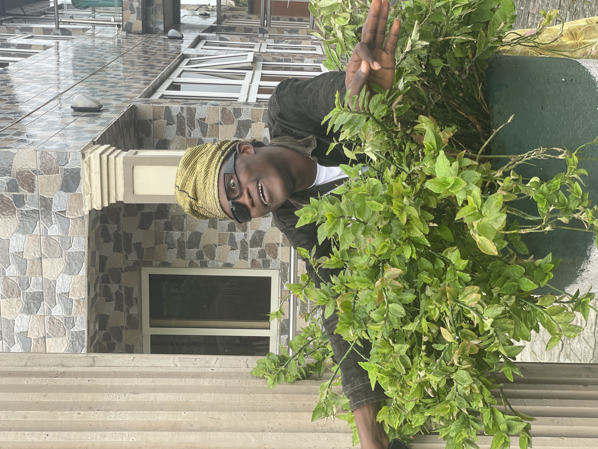 A young man, appearing to be in his late teens or early twenties, stands behind a large potted plant. He wears a gold patterned headwrap and dark sunglasses, along with a dark jacket over a white t-shirt. His mouth is open in a surprised expression, and he gazes upwards and to his left. His right arm is extended, with his hand resting on the edge of the planter, while his left arm is mostly obscured by the foliage.
The setting appears to be an exterior space, possibly a residential area or commercial building. Behind the man and the plant, a building with a facade of patterned tiles is visible. The tiles are in shades of beige, gray, and brown, arranged in a mosaic-like pattern. A large, fluted column is visible to the left of the frame, made of a light-colored material. The ground beneath the planter appears to be paved with light-colored stones. The lighting suggests it is daytime, with diffused natural light. The overall impression is a candid snapshot taken outdoors.
