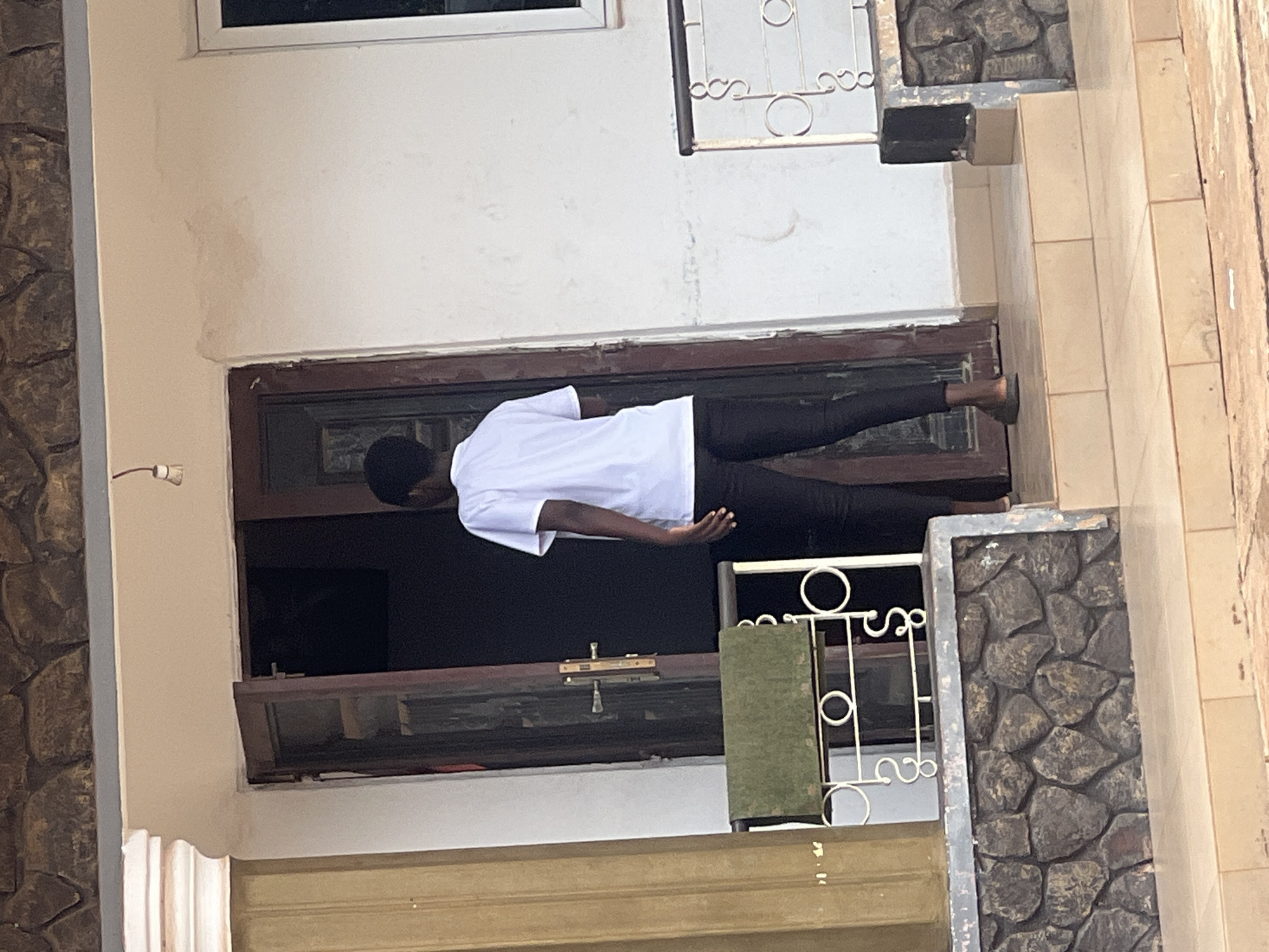 A person wearing a white t-shirt and black pants is standing on the doorstep of a building in Obosi, Nigeria. The building has a brown door frame, a window above the door, and a decorative wrought iron railing next to the doorway. The building is painted white and has a stone facade on one side of the doorway. The ground is tiled with light brown tiles. The person is facing away from the viewer.
