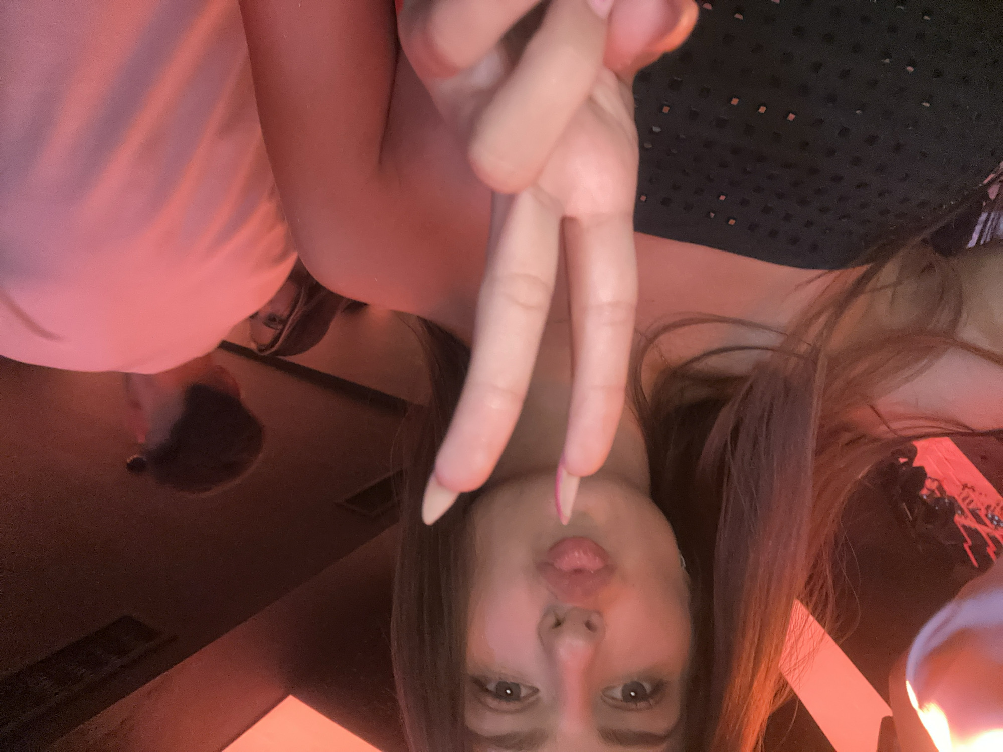 A young woman with long, brown hair and light skin is posing for a selfie. She is wearing a strapless black top. Her mouth is pursed in a kiss, and she is holding up two fingers in a peace sign. The background is dimly lit with red and orange hues, suggesting a club or bar. Other people are visible in the background, out of focus. The image appears to have been taken in Nesebar, Bulgaria, a location known for its nightlife.