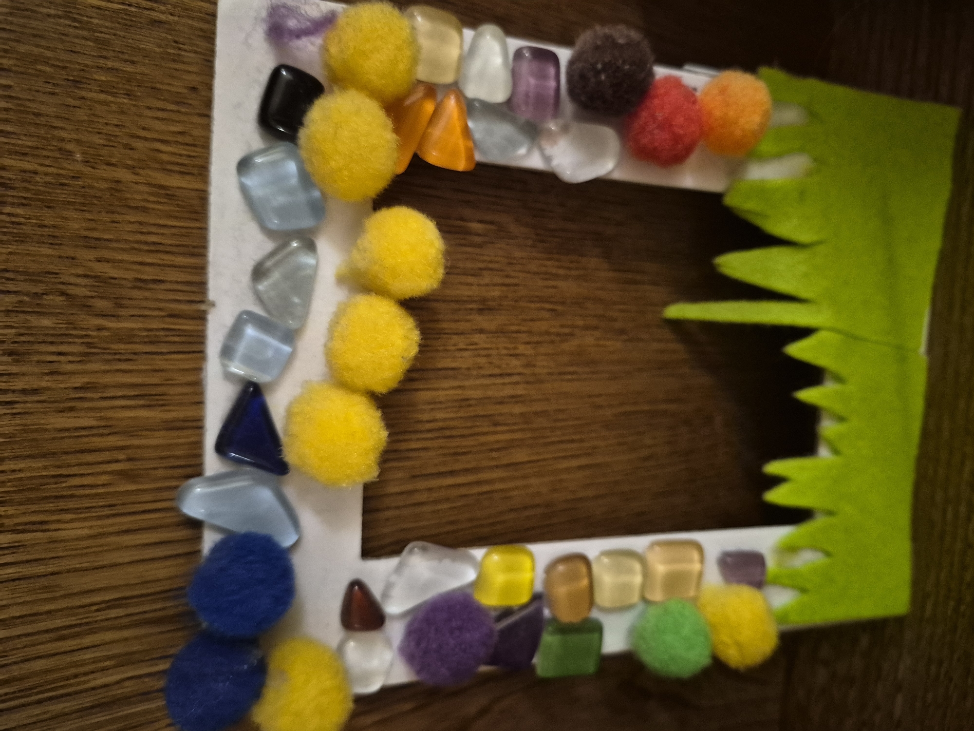 This is a vertical, eye-level, close-up shot of a handmade craft project, likely a picture frame, against a wooden background. The frame is made of white cardstock and is decorated with various colorful craft supplies. Along the top edge, there are blue pom-poms and light blue, dark blue, and clear glass gems. The right side of the frame features a border of clear and purple glass gems, with orange triangular gems interspersed, and yellow pom-poms. The left side of the frame is decorated with a mix of clear, green, yellow, and purple glass gems, as well as a purple pom-pom and a green pom-pom. The bottom of the frame is adorned with a strip of green felt cut to resemble grass blades. The background appears to be a wooden surface with a prominent grain pattern. There are no people or animals visible in the image. The lighting is somewhat dim, casting soft shadows.