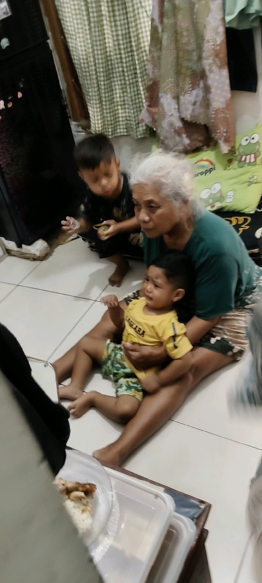 An elderly woman with white hair is sitting on the floor, holding a crying toddler on her lap. The toddler is wearing a yellow t-shirt with some text and camouflage shorts. Another young boy, wearing a black t-shirt, is standing near the woman and looking down. He is holding a small yellow object in his hand. They appear to be indoors, on a tiled floor. In the background, there is a dark object that could be a refrigerator or cabinet, and green and white checkered curtains. There are also pillows with cartoon frog designs on them. To the left of the frame, there is a clear plastic container with rice and some food. The scene suggests a domestic setting, possibly in a home in Mekarmukti, Indonesia.