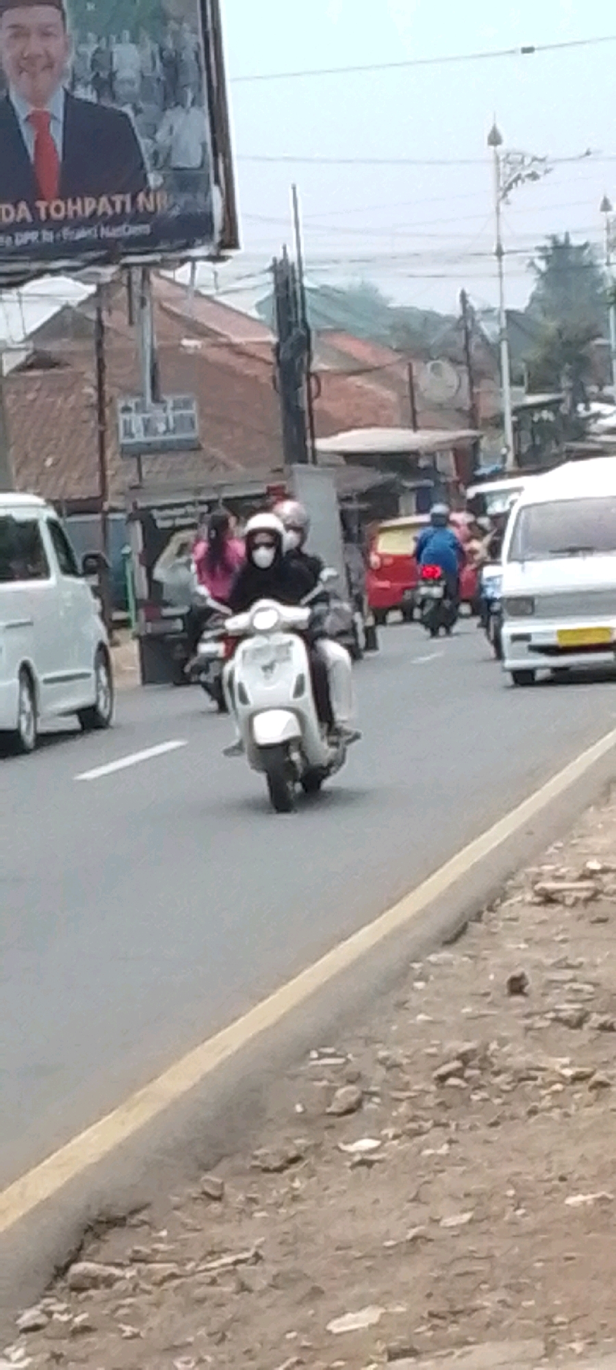 The image shows a street scene in Cilaku, Indonesia. A white scooter with two occupants wearing masks is in the foreground, traveling on a road. The rider is wearing a white helmet and the passenger is wearing a dark helmet. Behind them, a red car and a white van are visible, along with other vehicles and buildings. A billboard with a political figure is visible on the left side of the image. The environment appears to be an urban or semi-urban area with roads, buildings, and vegetation. The sky is overcast.