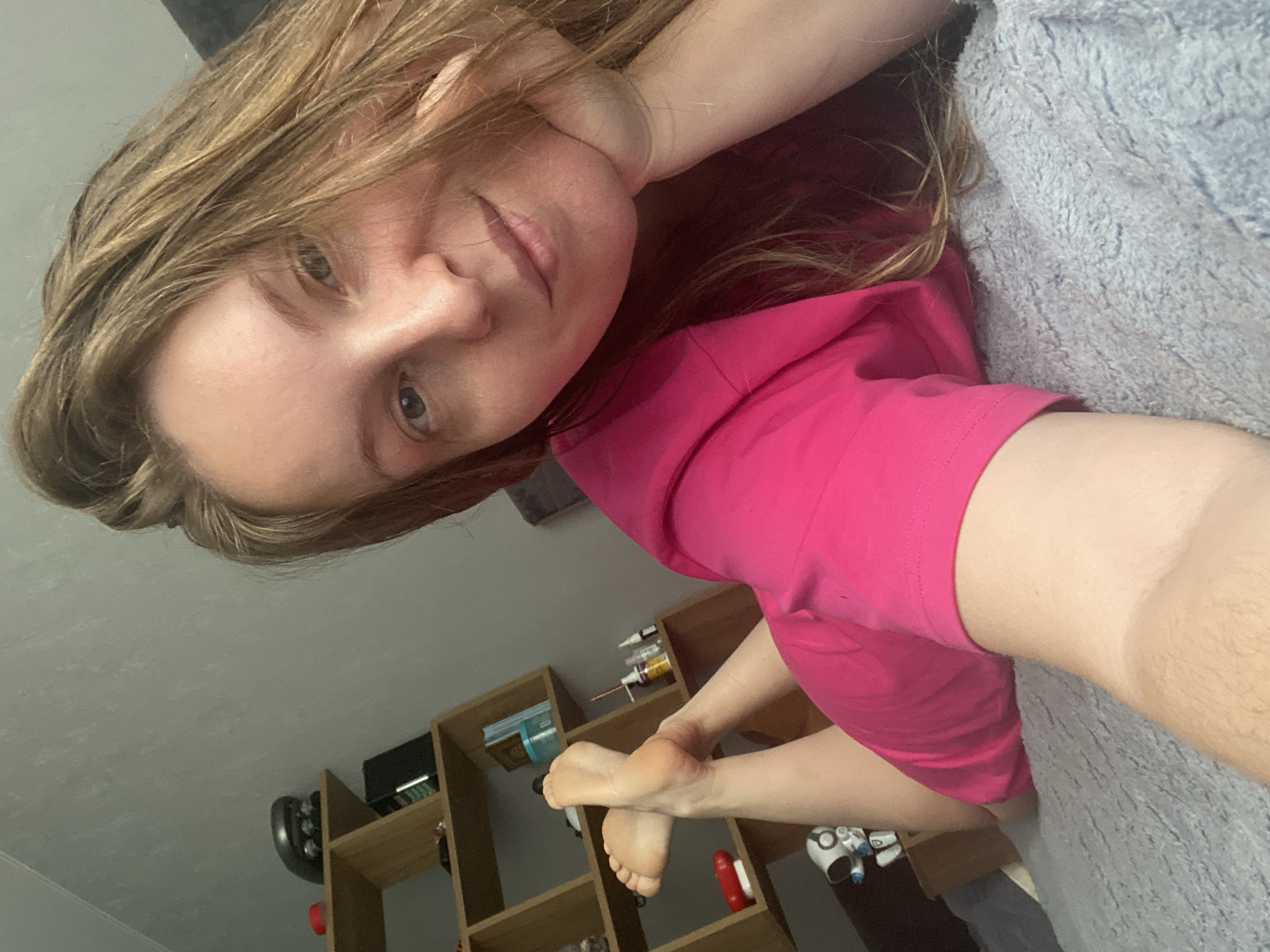 A woman with light brown hair and fair skin is lying on a gray blanket or sheet, wearing a pink t-shirt. Her head is resting on her hand. Her feet are visible in the lower part of the frame. Behind her, there is a light brown wooden bookshelf with various items, including books, bottles, and other objects. A dark gray wall is in the background. The location is unknown and cannot be determined based on the image.