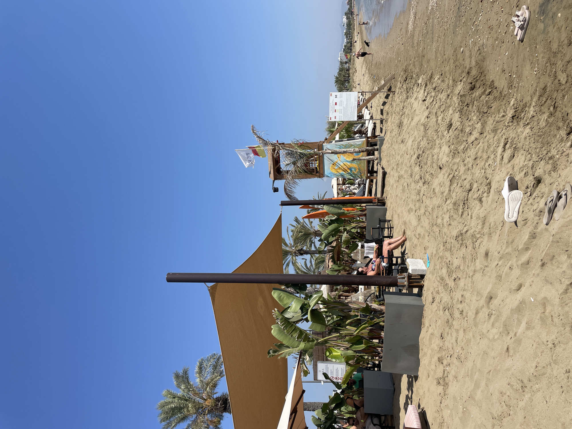 The image depicts a sunny beach scene in Trikomo, Northern Cyprus. The foreground is dominated by sandy beach with a few flip-flops and a small blue toy scattered on it. In the mid-ground, a shaded area is set up with tables and chairs, featuring lush green plants and palm trees. A woman is seated under the shade, facing away from the camera, wearing a bikini. Further back, a lifeguard tower stands tall, decorated with a painting of seahorses. A white flag with red lettering and a yellow and red flag are attached to the tower. Signs with information are also visible near the tower. The background shows the Mediterranean Sea, with a few people walking along the shore and in the water. Buildings are visible in the distance along the coastline, suggesting a populated area. The sky is clear and blue.