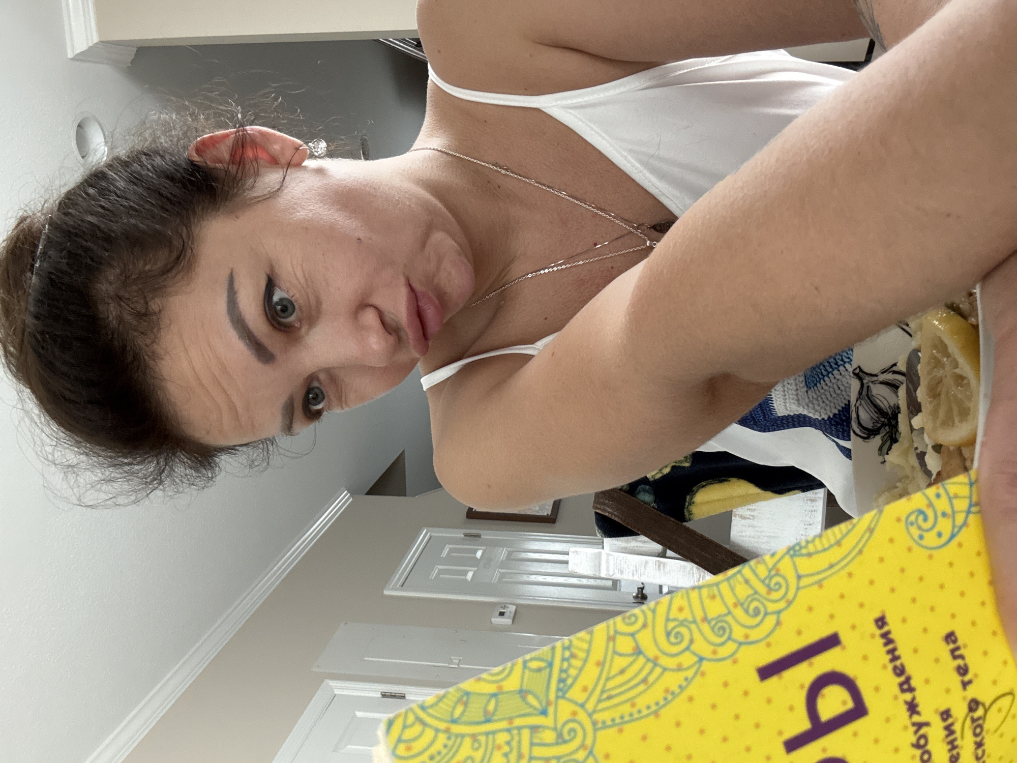 The image shows a woman with dark hair pulled back, wearing a white tank top and a silver necklace. She is looking at the camera with a surprised or inquisitive expression, her mouth slightly open. Her left arm is bent and resting in the foreground, partially obscuring a yellow object with a decorative pattern and some Cyrillic text. In the background, there are two white doors and a wall with molding. There is also a hint of food with lemon slices on a plate. The lighting suggests an indoor setting, possibly during the daytime. The overall mood is informal and candid. The visible text on the yellow object appears to be in Russian.