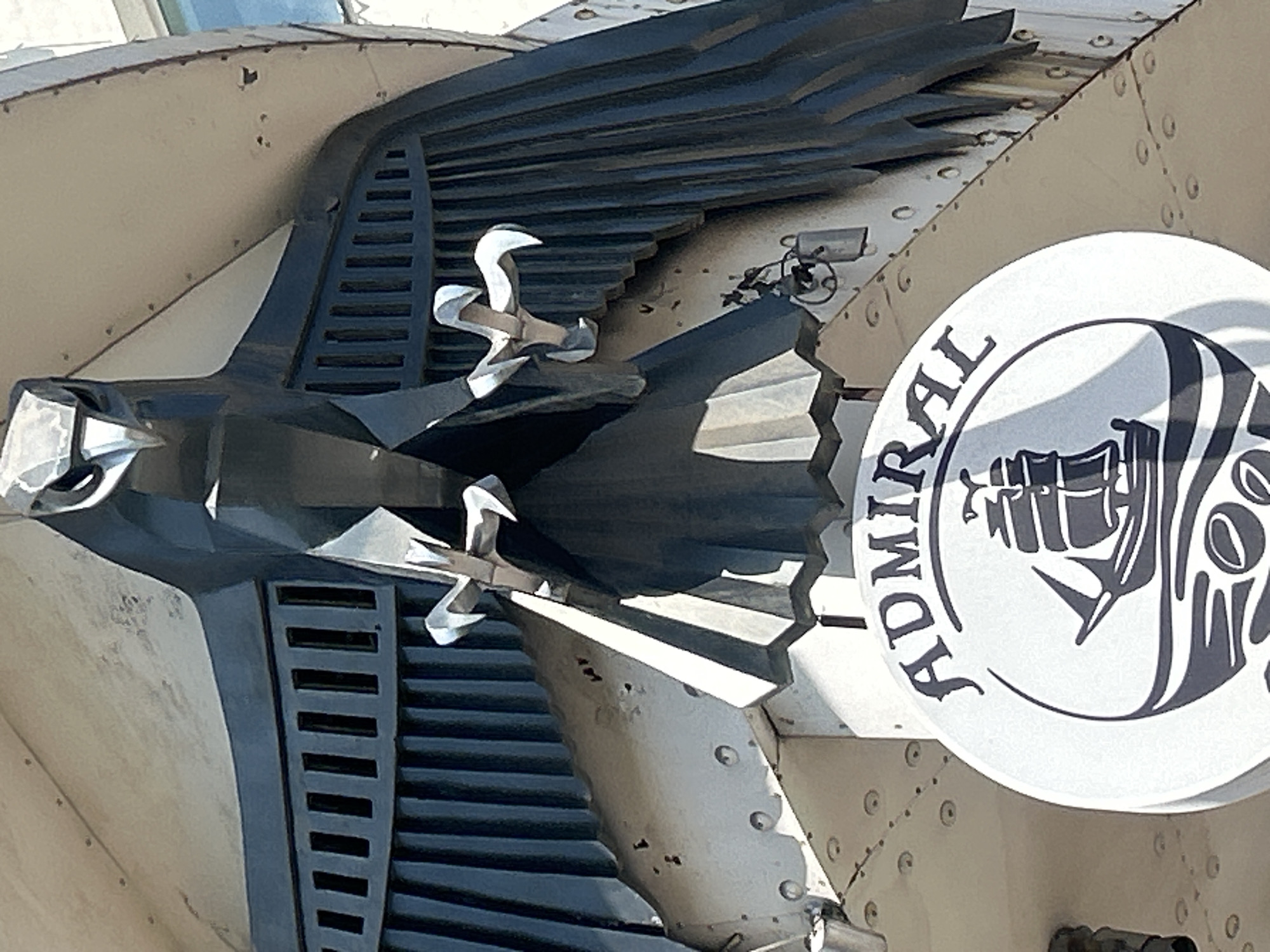 An external shot shows a stylized, dark metallic eagle sculpture with open wings, perched above a round white sign with a black silhouette of a sailing ship and the word "ADMIRAL". The eagle sculpture is mounted on a beige, riveted metallic structure, possibly part of a building facade. The sign appears to be for a business called "Admiral". The lighting suggests daytime. The scene is set in the city of Almaty, Kazakhstan.