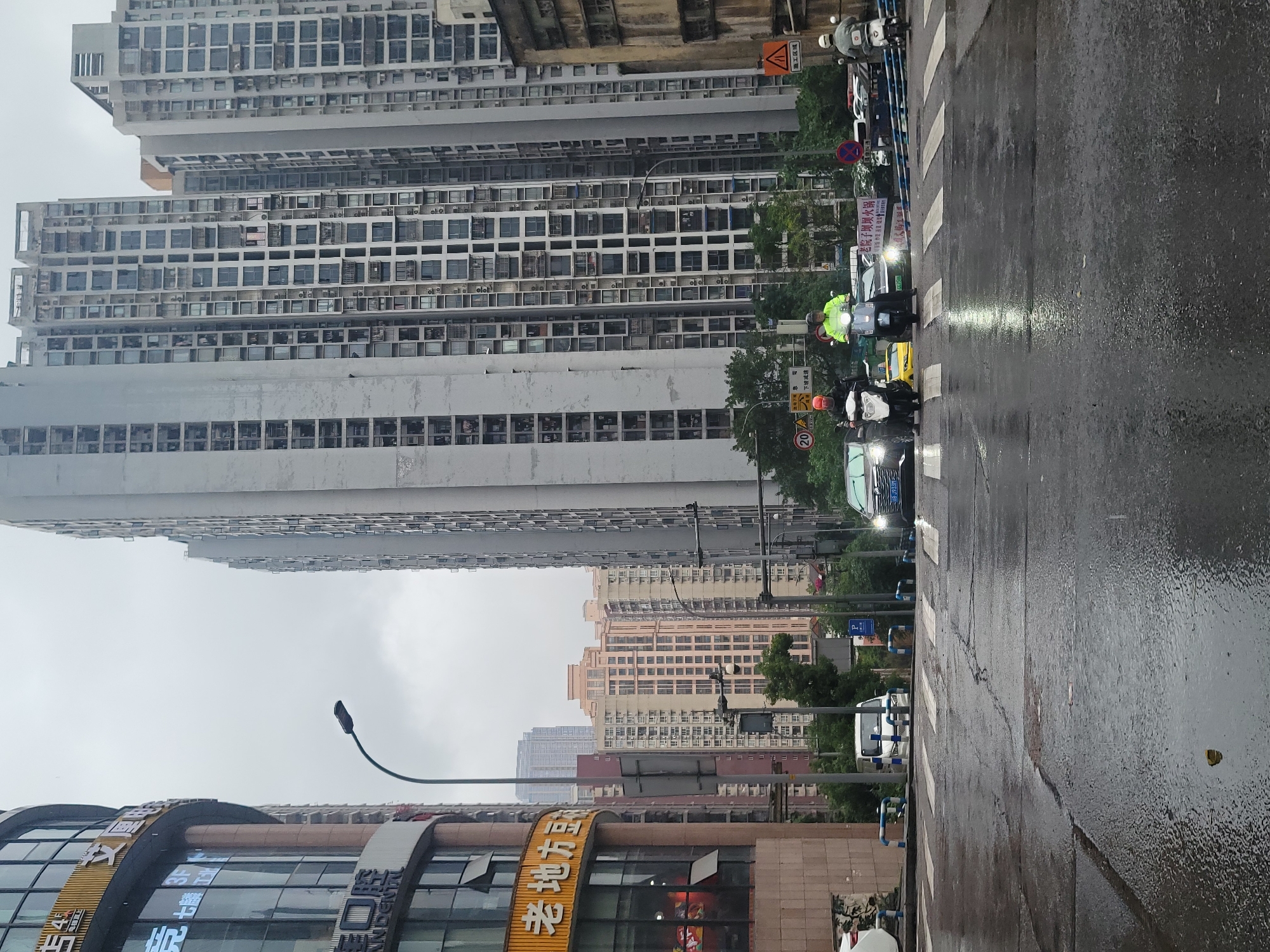 The image captures a dynamic urban street scene in Liangjiang, China, characterized by tall buildings and a wet, bustling road, suggesting recent rain. The perspective is from street level, looking down a thoroughfare lined with various structures.
In the background, a dense cluster of high-rise buildings dominates the skyline. These appear to be residential or mixed-use, with numerous windows and visible air conditioning units protruding from their facades. Some buildings are a drab grey, while others have a lighter, beige or light brown hue. The overcast sky indicates a cloudy day.
The foreground features a dark, wet asphalt road, reflecting the surrounding light and appearing slick from rain. White crosswalk markings are visible across the street. Several motorbikes and scooters are present on the road; some have their headlights illuminated, casting bright beams. One person on a scooter wears a bright yellow-green jacket, standing out against the muted tones of the environment. Cars are also visible in traffic.
On the left side of the image, closer to the viewer, is a commercial building with prominent signage. Visible text includes "口腔 ANDENIAL" (likely indicating a dental clinic), "老地方豆", and "3F". Further along the street, other signs include "老院子坝坝火锅" (Old Courtyard Babba Hot Pot), "施工区域" (Construction Area), "下坡减速" (Slow Down on Downgrade), and a speed limit sign showing "20". Streetlights and sparse trees are also part of the urban landscape.
The overall impression is an active, modern city environment, experiencing wet weather conditions.