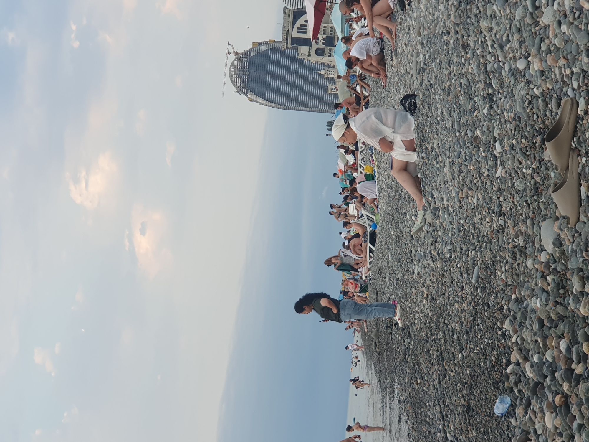 The image depicts a crowded pebble beach in Batumi, Georgia, with the distinctive curved skyscraper under construction, likely the Hilton Batumi, in the background. The sky is overcast with patches of bright clouds. Numerous people are present, some wading in the calm sea, while others are relaxing on lounge chairs or sitting directly on the pebbles. A woman in a dark t-shirt and jeans stands in the foreground, looking at her phone. Further back, a person in white clothing with a headscarf is seated on a lounge chair. Beach umbrellas are scattered across the scene. A plastic water bottle lies on the pebbles in the lower left corner, and a pair of sandals are visible in the bottom center. The overall impression is of a typical summer day at a popular seaside resort.