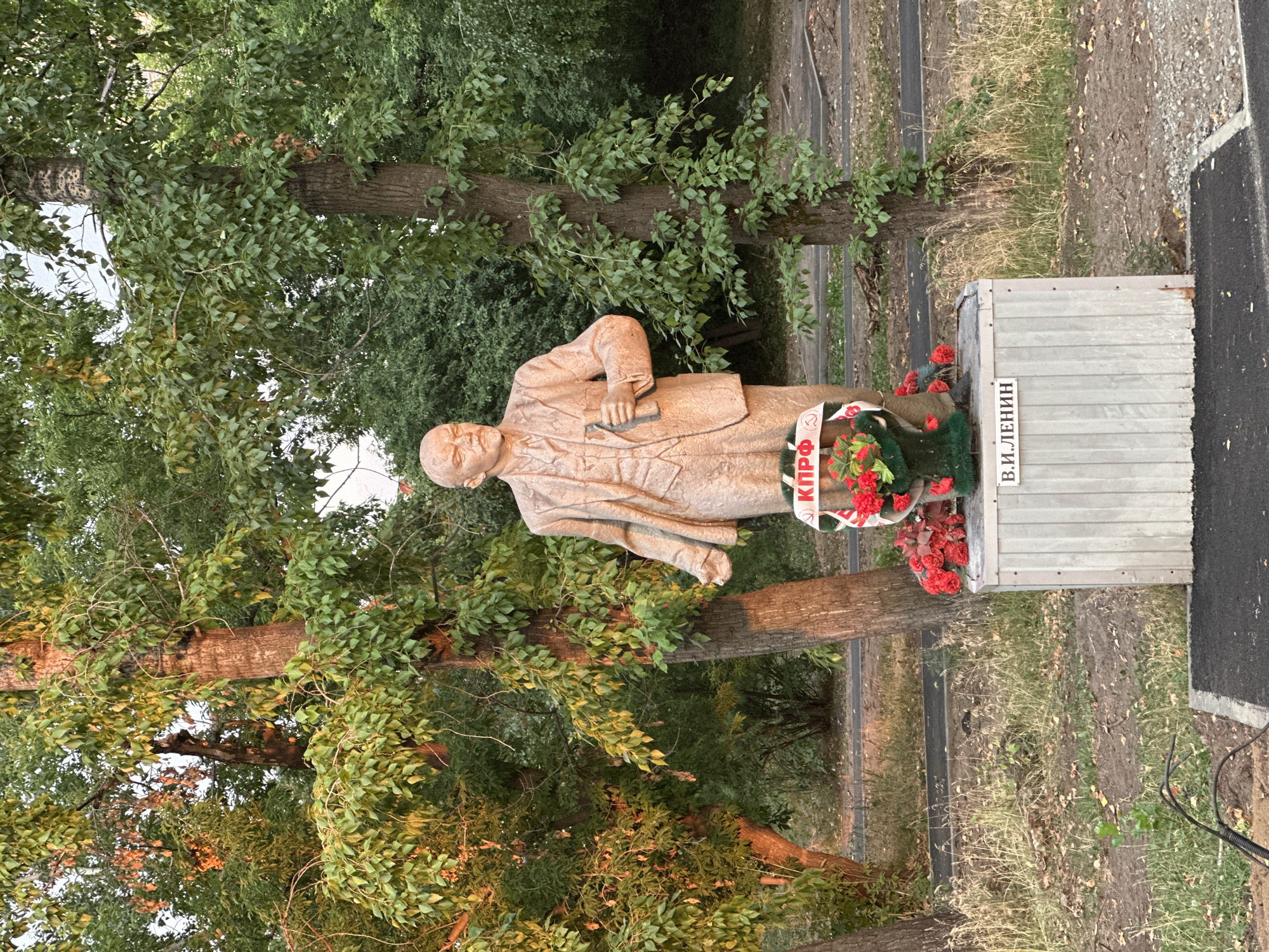 The image shows a statue of Vladimir Lenin in Biysk, Russia. The statue is made of bronze and depicts Lenin standing with a briefcase in his hand. He is wearing a suit and tie. The statue is located in a park, surrounded by trees. There are also some flowers placed at the base of the statue. A wreath with red flowers is around Lenin's neck. On the base of the statue, there is text that reads "В.И. ЛЕНИН" (V.I. Lenin). The photo appears to have been taken during the day, with overcast skies. The overall mood of the image is somber, reflecting the historical significance of Lenin and his legacy.