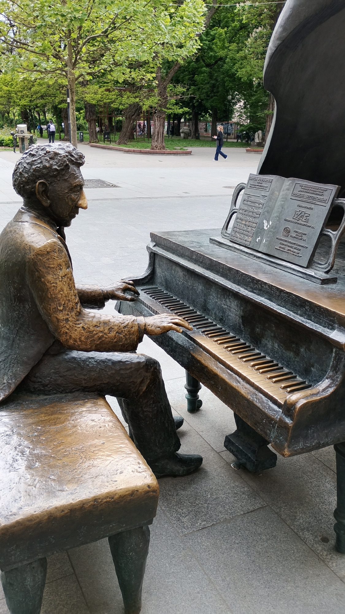 The image shows a bronze sculpture of Arthur Rubinstein playing a grand piano. The sculpture is located in a public square with trees and people in the background, likely in the city of Łódź, Poland. The sculpture depicts Arthur Rubinstein seated at a grand piano, dressed in a suit, with a focused expression as his hands rest on the piano keys. Sheet music is displayed on the piano's music rest. The environment includes a paved square with trees and other pedestrians in the distance. The scene is outdoors and appears to be during the daytime.