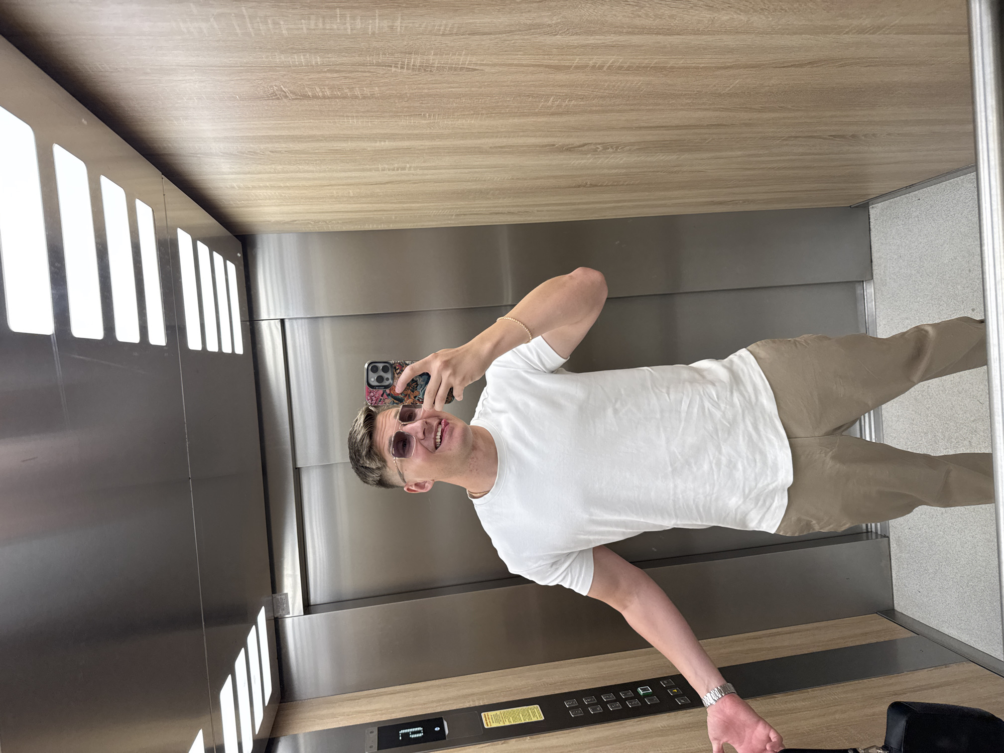 A young man is taking a selfie in an elevator. He is wearing a white t-shirt, khaki pants, and sunglasses. He has a watch on his left wrist and a bracelet on his right wrist. He is holding a smartphone with a patterned case. The elevator has wood-paneled walls and a stainless steel control panel. The elevator also has a panel of lights on the left side. It is impossible to confirm the location is in Plovdiv, Bulgaria, based on the image alone.