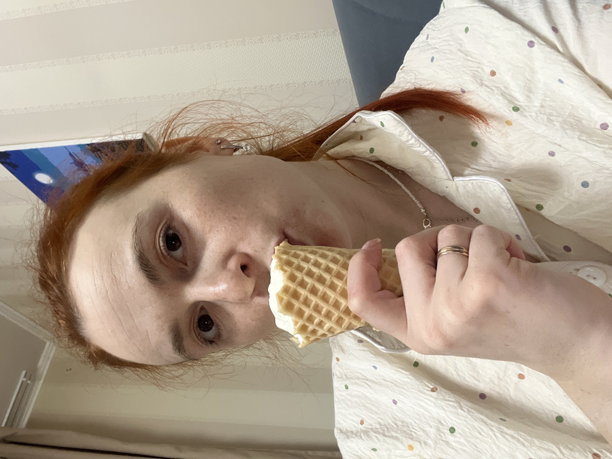 A pale-skinned individual with reddish hair is consuming an ice cream cone. The ice cream is white and held in a waffle cone. The individual is wearing a light-colored shirt with multi-colored polka dots and a small silver necklace. A gold ring is visible on a finger. The background includes a striped wall and a glimpse of a framed artwork. The location is Chaikovsky, Russia.
