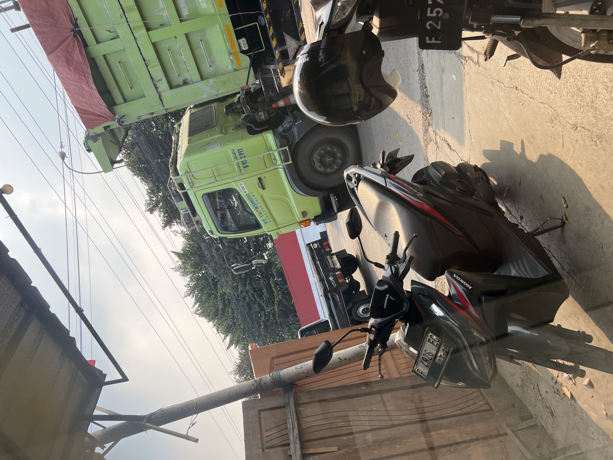 The image depicts an outdoor scene in Citeureup, Indonesia. In the foreground, two motorcycles are parked. The one on the left is black with red accents and has a license plate reading "F 4826 EG" with an expiration date of "07-28". A black helmet rests on the handlebars of the motorcycle on the right, which has a license plate starting with "F 257". Behind the motorcycles, a black pickup truck with a white logo that says "CARRY" is visible. Further in the background, a large green dump truck is parked. The truck has text on its side, including "PT. TRANSIINDO" and "PERKASA PRIMA". A red tarp covers the bed of the dump truck. The scene is set against a backdrop of trees and utility wires. A building with a red roof is partially visible behind the pickup truck. Wooden panels are stacked next to a concrete pillar supporting an awning. The lighting suggests it is daytime, with sunlight casting shadows on the ground.