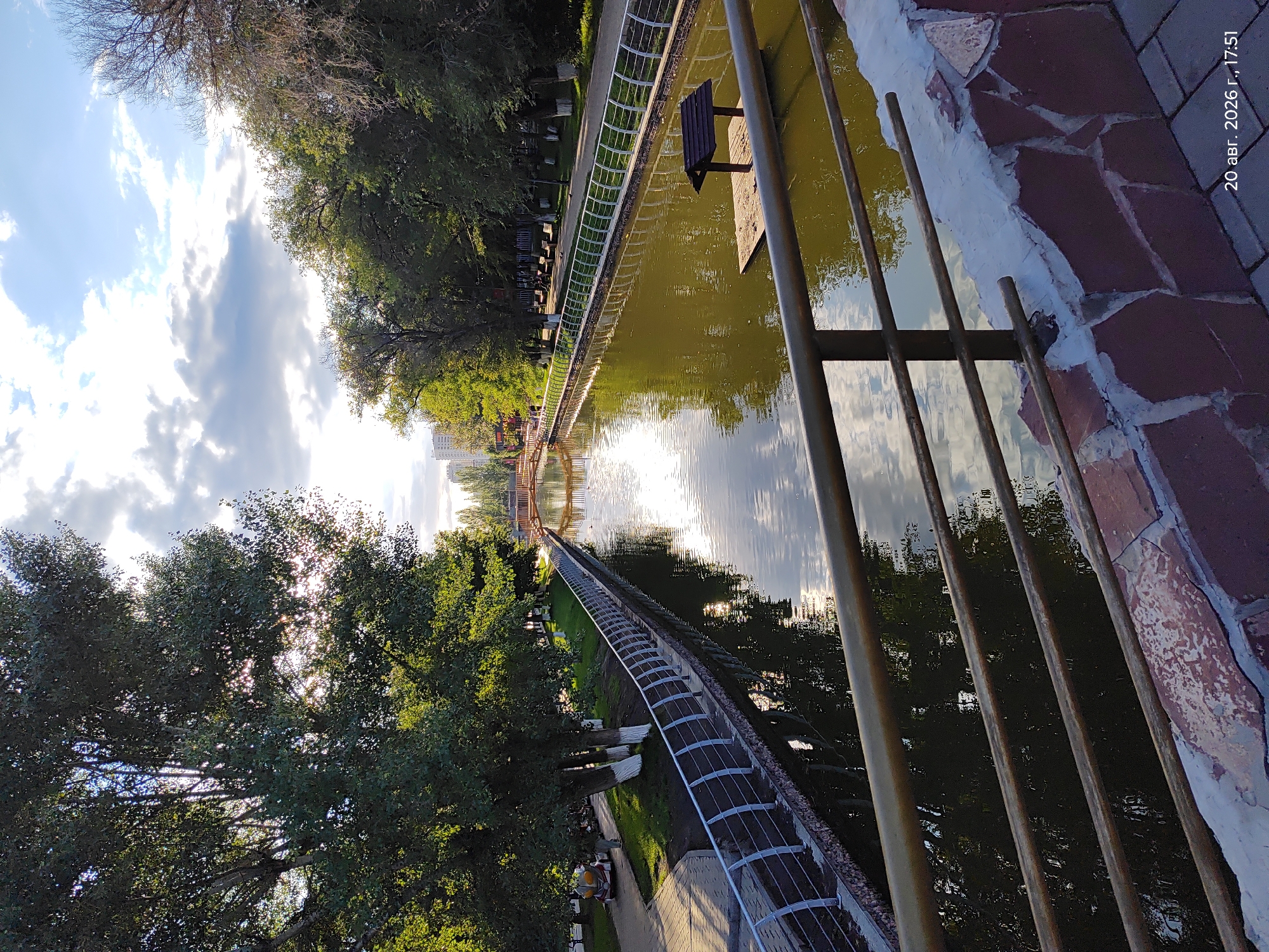 The image shows a canal with a walkway on the left and trees lining the right bank. A railing made of metal bars and a stone wall runs along the foreground of the canal. The water reflects the sky and trees. In the distance, a pedestrian bridge crosses the canal, and beyond that, more trees and buildings are visible. A small wooden structure on a floating platform sits in the canal on the right. The image is dated "20 авг. 2026 г., 17:51", which translates to "August 20, 2026, 17:51". Based on the general appearance of the architecture and vegetation, this scene is likely located in Astana, Kazakhstan, potentially in a park or public area with a waterway.