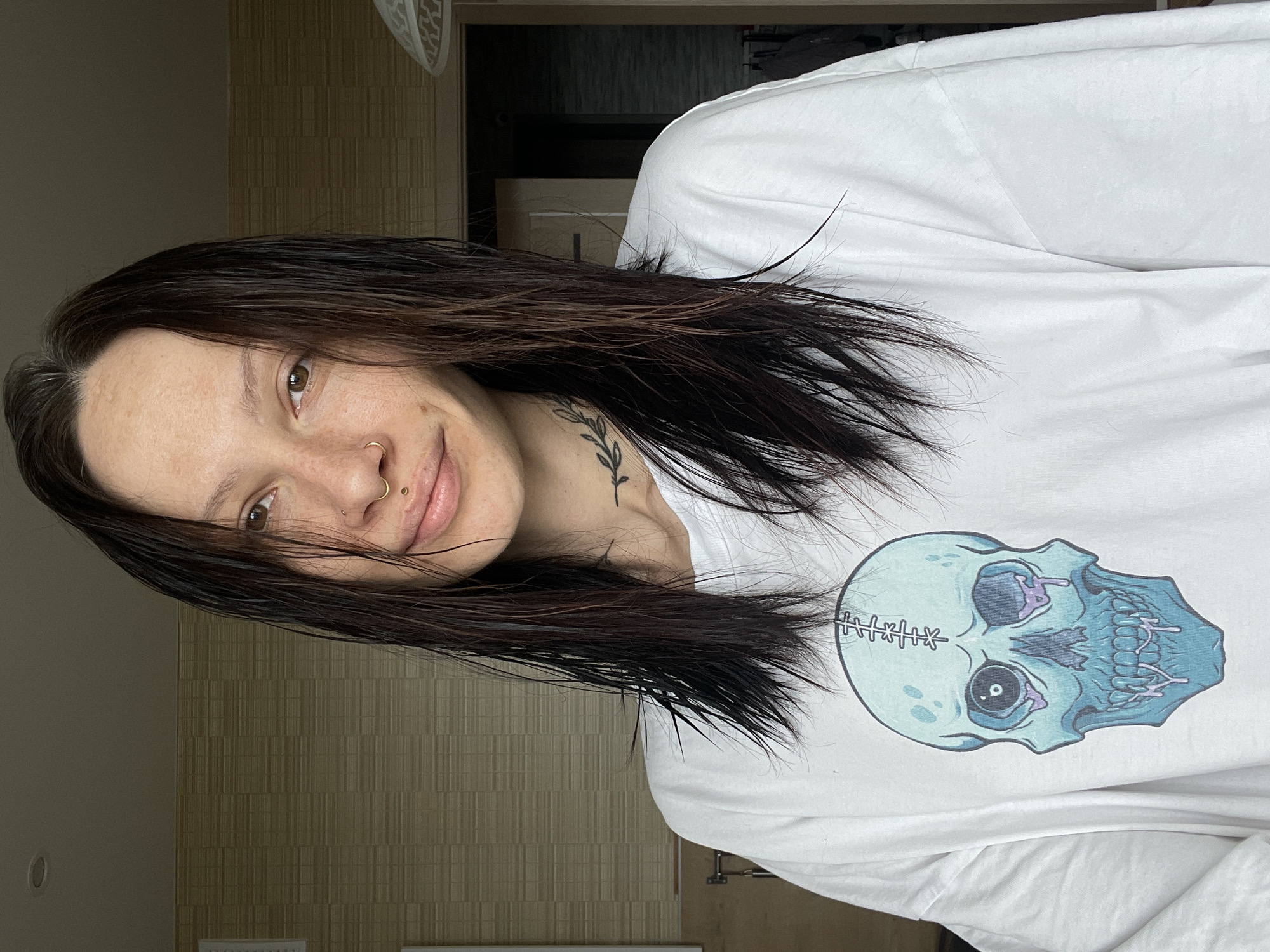 A young woman with dark brown hair and a nose ring is posing for a photo. She is wearing a white long-sleeved shirt with a graphic of a skull on the front. A small tattoo of a sprig of leaves is visible on her neck. The background appears to be a room with textured walls and a glimpse of an open doorway. The location of the photo is indeterminable, and there is no clear indication that this image was taken in Izhevsk, Russia.