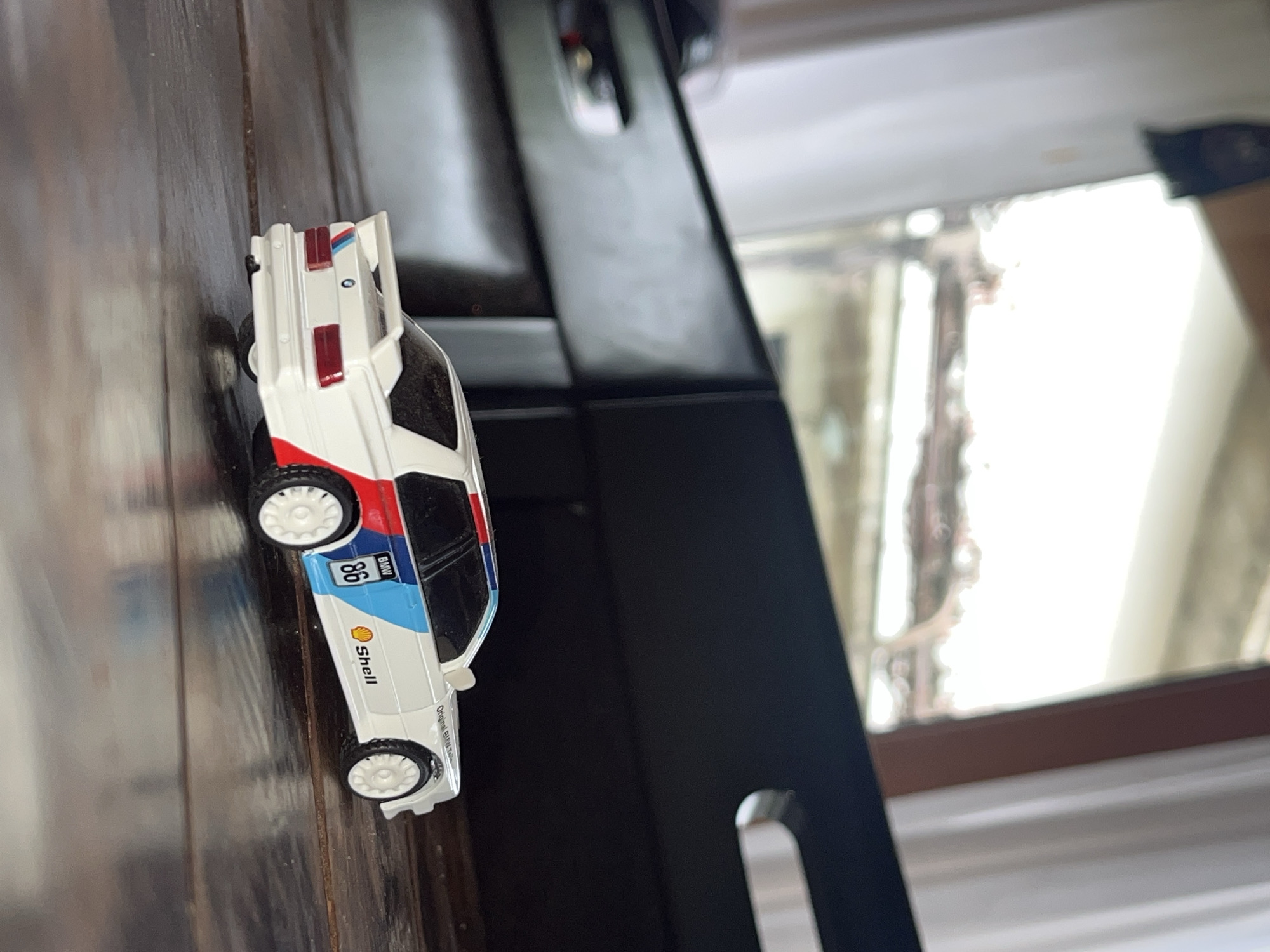 The image shows a white toy car with red and blue stripes and "Shell" branding, lying on a dark wooden surface. The car has the number "86" on its side. Behind the car, there is a black metal structure with rounded edges. In the background, there is a window covered with sheer curtains. The location is not discernible from the image. There are no people visible.