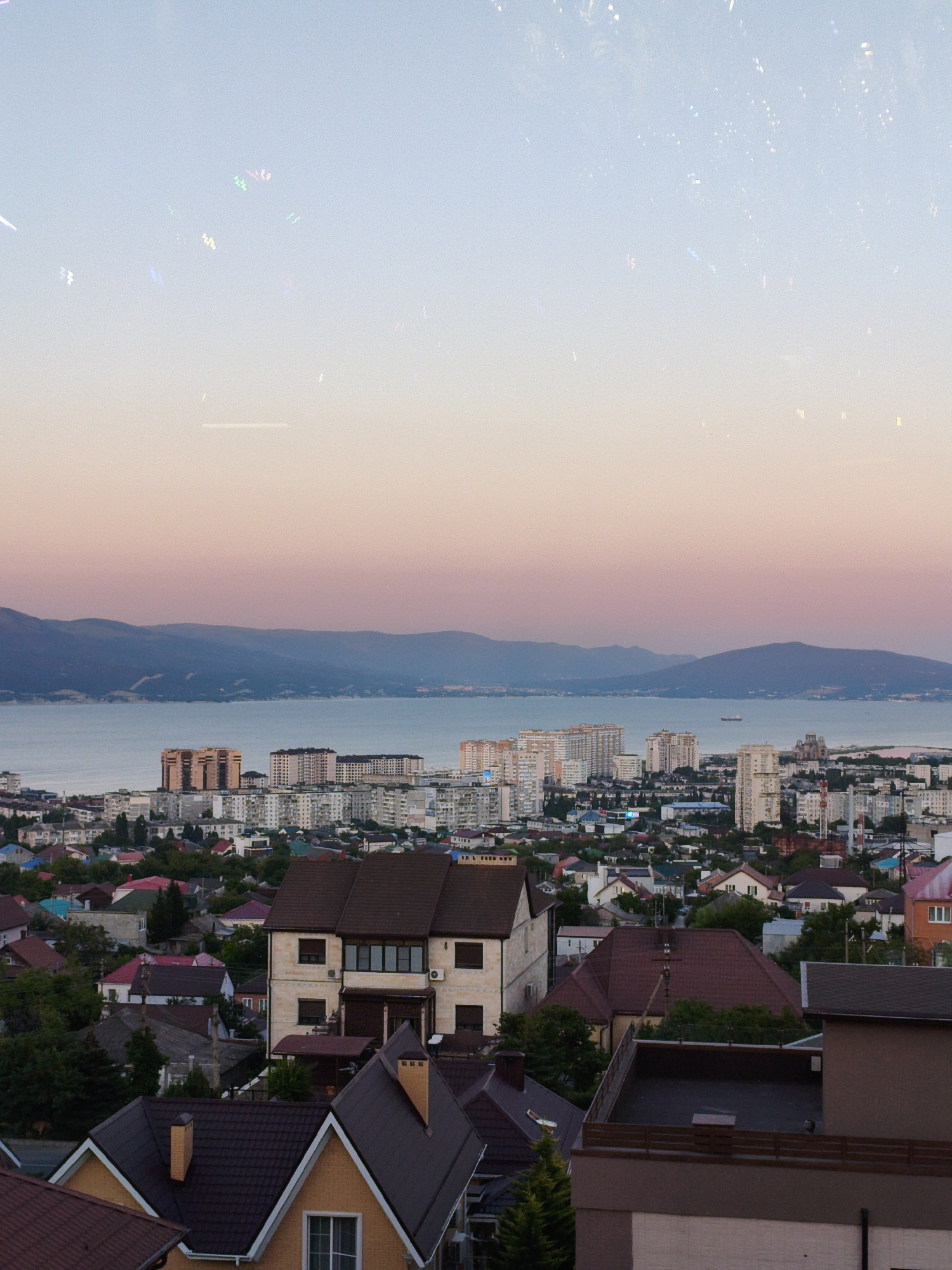 The image depicts an aerial view of a city situated on a coast, with a body of water and mountains in the background. The sky transitions from a pale blue at the top to a soft pinkish hue near the horizon, suggesting either dawn or dusk. Several buildings, ranging from multi-story apartment complexes to smaller houses with pitched roofs, are visible in the foreground and midground. The architectural style appears to be primarily modern, with some older residential structures. A body of water, likely a sea or large lake, separates the city from a range of low, rolling mountains. The overall scene conveys a sense of urban development along a natural landscape. There are no people or specific actions depicted in the image.