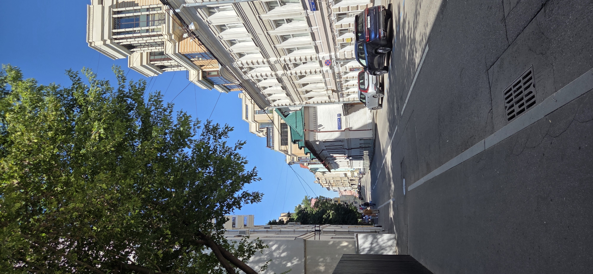 The image depicts a street scene in Moscow, Russia. The perspective is from a sidewalk looking down a street lined with buildings on both sides. A large tree with green foliage overhangs the left side of the image, casting shadows on the sidewalk.
On the right side of the street, several multi-story buildings are visible. The closest building on the right has a light-colored facade with numerous windows and ornate architectural details. Further down the street, other buildings of varying heights and architectural styles can be seen, including one with a green roof and another modern building with a light facade and large windows.
Two cars are parked on the right side of the street. A dark-colored SUV is closer to the foreground, and a lighter-colored sedan is parked in front of it. In the distance, several other vehicles are present on the road.
On the left side of the street, a darker building with a black fence is visible in the foreground. Further down, several pedestrians are walking on the sidewalk. The street itself is paved and has white lane markings. A storm drain grate is visible in the foreground on the asphalt. The sky is clear and blue.