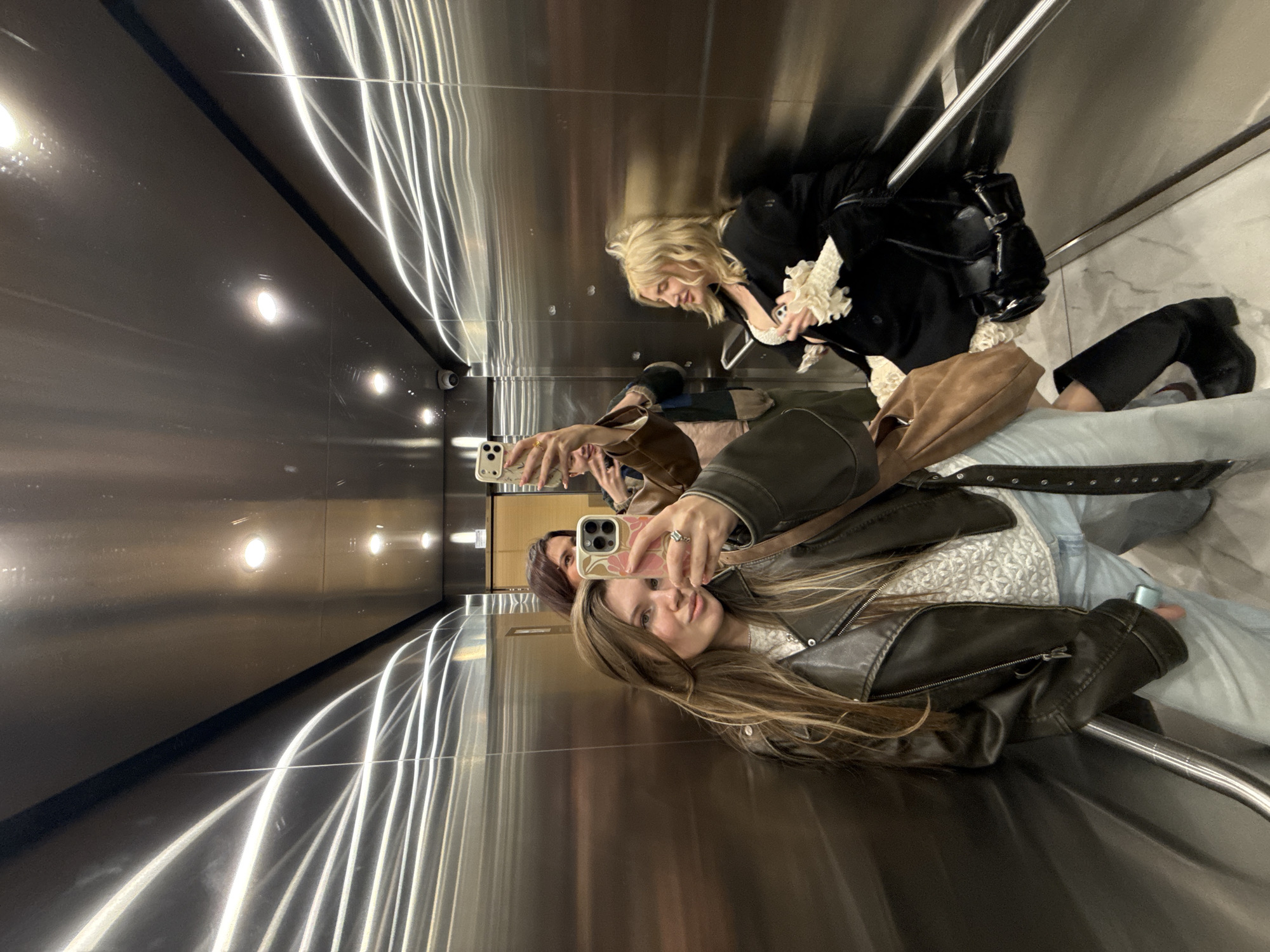 Three women are taking selfies in an elevator. One woman is in the foreground wearing a leather jacket. Another woman is behind her, and a third woman is behind the second. The elevator has brushed metal walls with light reflections. The location is likely within the city of Belgrade, Serbia, based on visual metadata hints.