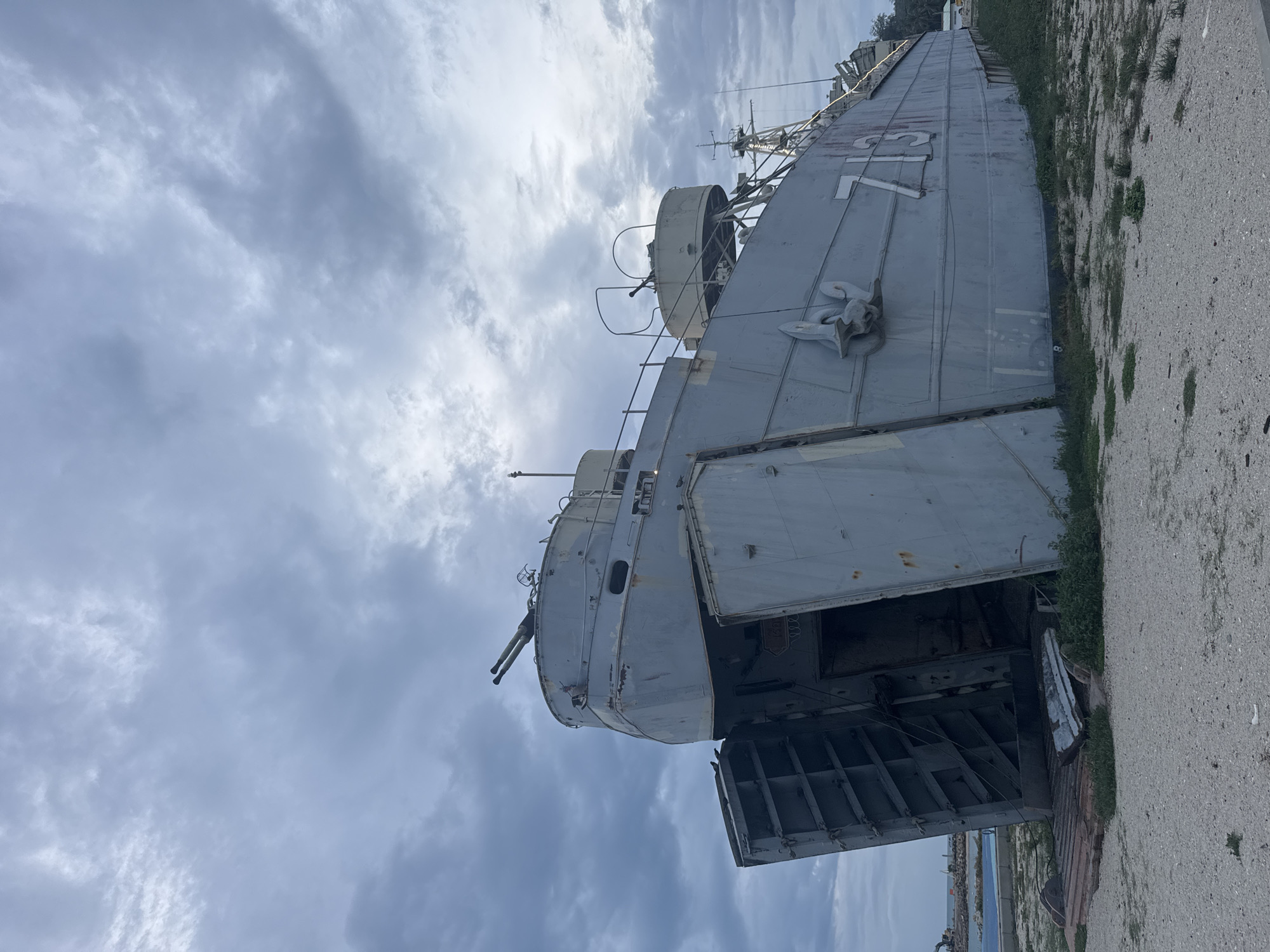 The image shows the LCT-770, a decommissioned U.S. Navy landing craft tank, displayed on land in Thong Sala, Thailand. The vessel is painted gray and appears weathered with rust and peeling paint. The hull number "770" is visible near the bow. The front loading ramp is lowered, revealing the interior. A twin-barreled gun is mounted on the forward superstructure. The sky is overcast with gray clouds. The ship sits on a concrete surface adjacent to a patch of grass and some unidentified structures in the background near the water.