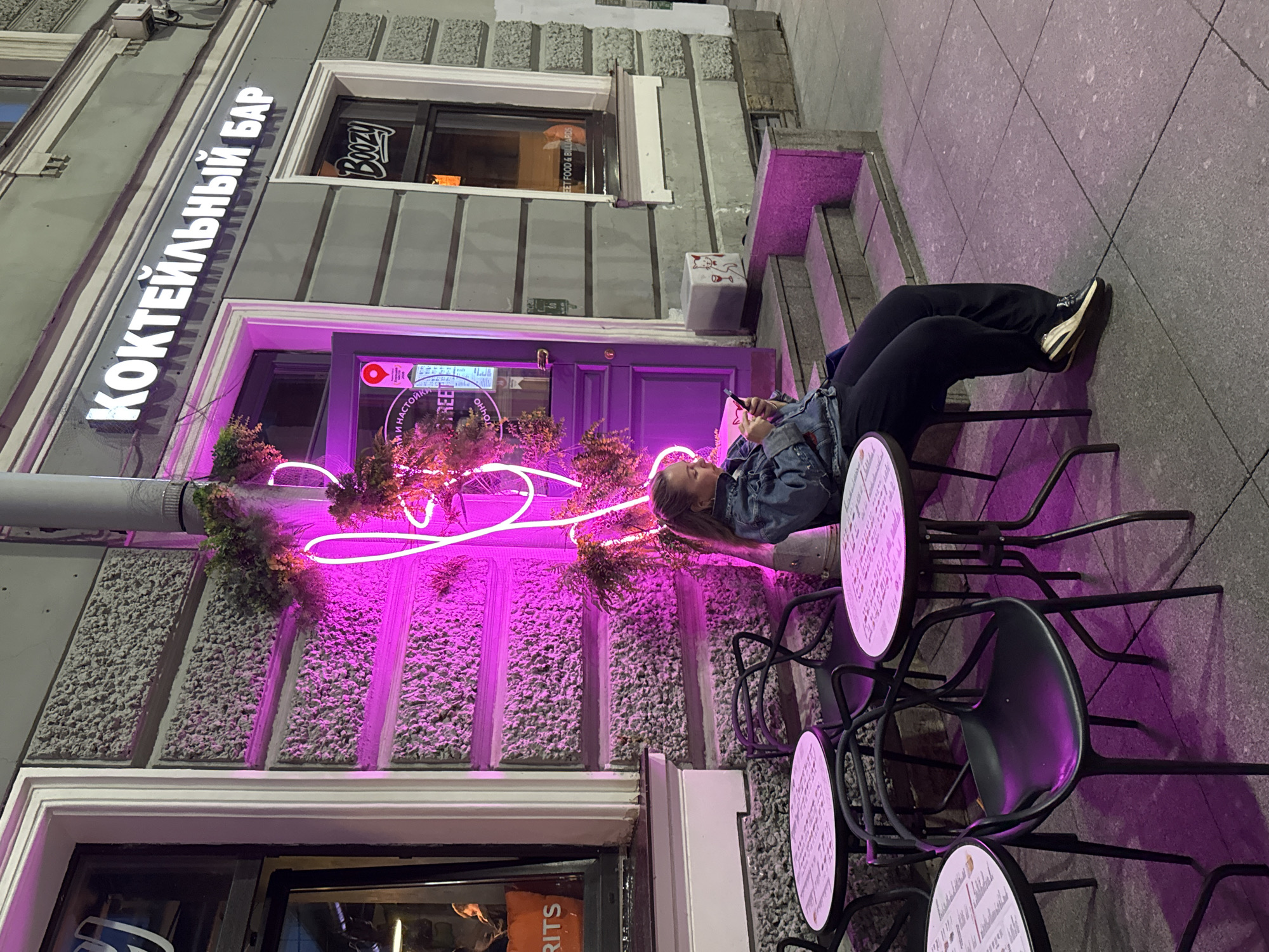 A young woman with blonde hair pulled back in a ponytail is sitting at an outdoor table at a cafe at night. She is wearing a denim jacket and black pants and is looking down at her phone. The cafe has neon pink lighting, and there are tables and chairs set up on the sidewalk. The sign above the cafe reads "КОКТЕЙЛЬНЫЙ БАР," which translates to "Cocktail Bar." The cafe appears to be located in the city of Saint Petersburg, Russia, as indicated by the Cyrillic text on the sign. There are other tables and chairs visible, and a staircase leading up to the entrance of the establishment. The overall atmosphere is urban and modern, with a hint of neon-lit ambiance.