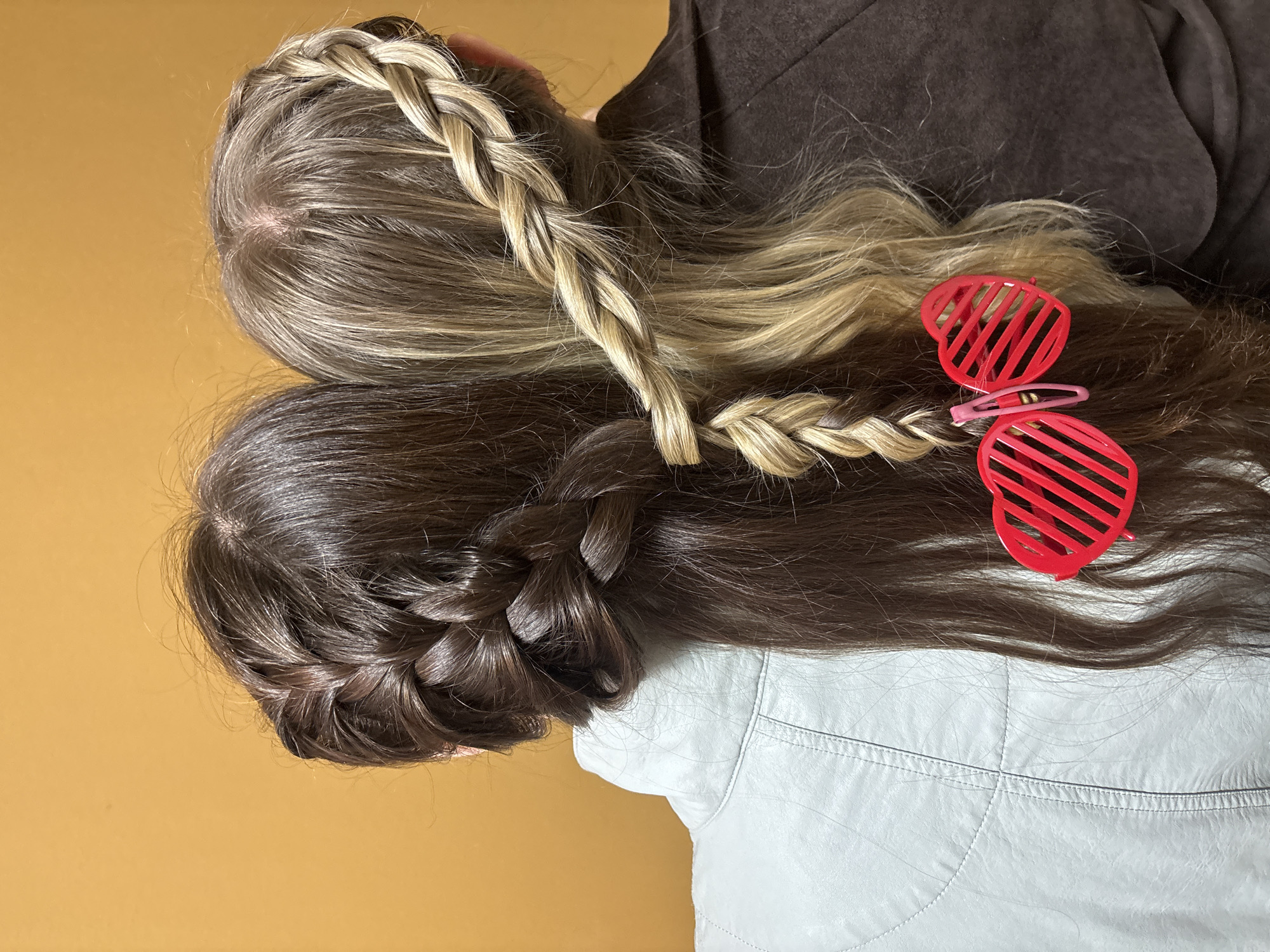 The image displays a close-up, overhead view of the backs of two individuals, likely taken indoors in Chaikovsky, Russia, against a plain, solid mustard-yellow background.
The subjects are two people, distinguishable by their hair color and style. The person on the upper left has long, predominantly blonde or light brown hair with subtle highlights, styled with a prominent, tightly woven blonde braid running diagonally across their head. They are wearing a dark brown jacket or garment. The second person, positioned to the lower right, has long, dark brown hair, also styled with a thick, intricate braid on the side of their head. This individual is wearing a light-colored, possibly white or light grey, jacket with visible stitching detail, suggesting a leather or similar fabric.
A central element of the image is the interplay of their hairstyles and accessories. A thinner blonde braid from the first person's hair appears to intertwine with a dark brown braid from the second person's hair. Adorning the hair near this intersection are two bright red, slatted, heart-shaped hair clips, along with a smaller pink hair clip. These accessories serve as a decorative focal point.
The activity appears to be a posed photograph, showcasing the detailed braided hairstyles and the use of the heart-shaped hair clips. The lighting is bright and even, casting no harsh shadows, suggesting an indoor setting with good ambient light. There is no visible text in the image. No specific time of day or emotions are discernible.