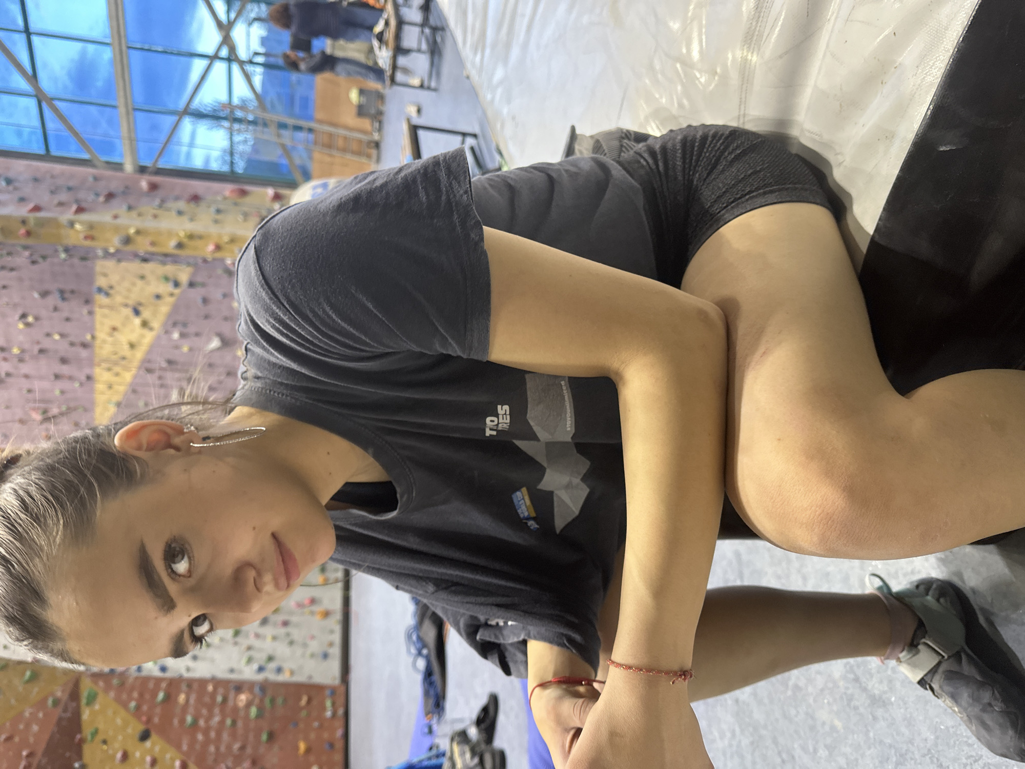 This image captures a young woman in an indoor climbing or bouldering gym, located in Sofia, Bulgaria. She is positioned in the foreground, seated or leaning on a grey and white padded crash mat, looking directly at the camera with a neutral expression.
She is wearing a dark, short-sleeved athletic t-shirt, upon which text can be seen, including "TIO RES" and "#stayyouunlimited." She also wears dark athletic shorts, and a red string bracelet is visible on her left wrist. A climbing shoe is worn on her right foot. Her hair appears to be pulled back, and she has distinctive long, hoop-style earrings.
The background reveals a vibrant climbing environment with multiple climbing walls adorned with numerous colorful holds of various shapes and sizes. Other individuals are visible in the distance, engaged in climbing activities. The gym's architecture includes a prominent glass roof or wall section, allowing natural light to enter and reflecting parts of the interior. Additional climbing equipment, such as ropes and shoes, can be seen on the floor. The scene suggests a moment of rest or observation during a climbing session in an active and modern recreational facility.