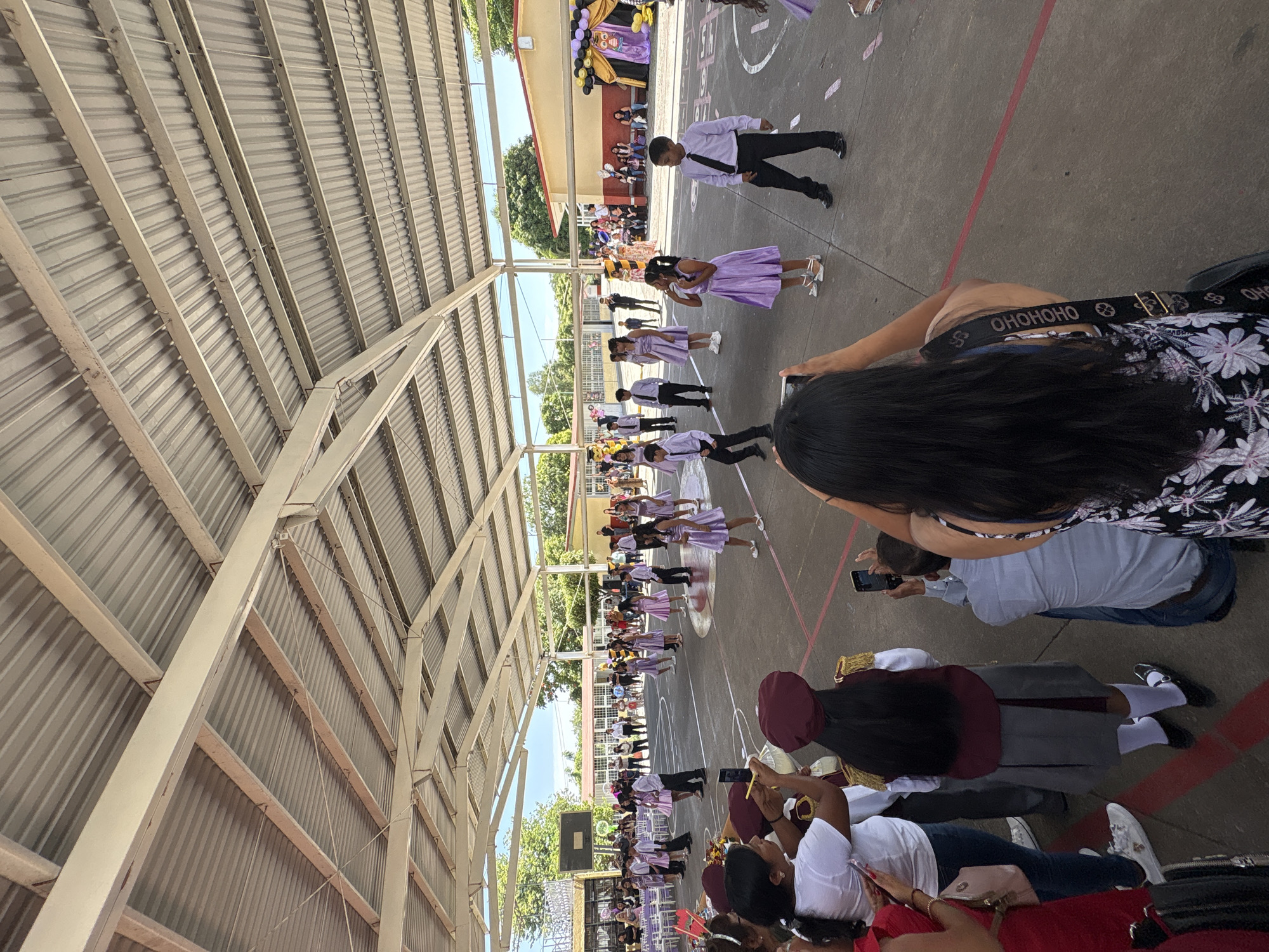 This is a full-shot, eye-level, outdoor photograph taken in Tejalpa, Mexico, depicting a school event under a large, open-air canopy. The canopy has a corrugated metal roof supported by a white metal frame. In the foreground, several people are visible from behind, facing a group of children performing in the middle ground.
The performers, composed of boys and girls, are dressed in formal attire. The girls are wearing light purple dresses and white shoes, while the boys are in white shirts, black ties, black pants, and black shoes. They are arranged in formation on a paved, outdoor court marked with red and white lines, possibly a basketball court. Some of the children appear to be dancing or marching.
In the foreground, spectators are holding up cell phones to take pictures or record the event. One woman in the immediate foreground is wearing a white t-shirt and has a maroon and gold colored uniform on her back. Another woman is wearing a black and white floral top and has a black bag with a patterned strap.
In the background, beyond the performance area, there are buildings, trees, and a basketball hoop. To the right, a decorated archway with balloons in black and gold suggests a celebratory atmosphere. The overall scene appears to be a school performance or graduation ceremony.