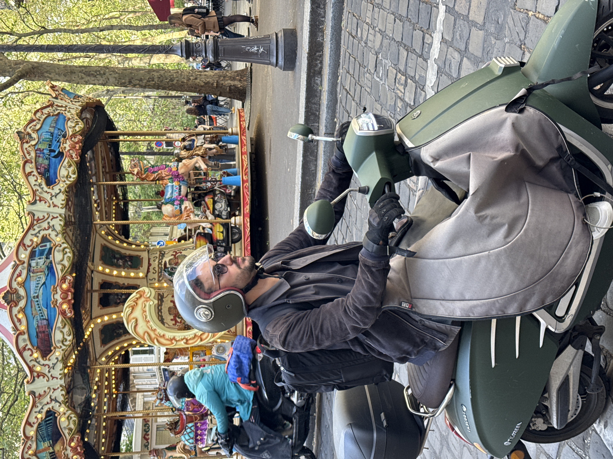 The image displays a lively street scene, rotated 90 degrees clockwise, set in what appears to be Paris, France, on a bright, sunny day. In the immediate foreground, a man is seated on a dark green scooter. He wears a grey helmet with a clear visor, sunglasses, a dark jacket, and black gloves. A black backpack is strapped to his back, and a grey protective cover drapes over the scooter's front, covering his legs. He appears to be stopped, looking towards his right.
Behind the man, a traditional, ornate carousel with multiple lights and painted panels, featuring a cityscape, is prominent. Several horse figures are visible on the carousel, and people can be seen around it, some appearing to ride. Mature trees with vibrant light green foliage line the background, suggesting spring or early summer. The street surface transitions from cobblestone pavement on the right to an asphalt road further left. A classic dark lamppost stands near the trees. Another person on a scooter, wearing a light blue jacket, is partially visible behind the main subject.
Visible text includes "PLUS" on the grey leg cover of the scooter, and "TILSCOOT" and "TILGREEN" (partially obscured) are written on the scooter's rear fender. "TILSCOOT" is also faintly visible on one of the side mirrors. The scene captures a moment of urban activity and recreation.
