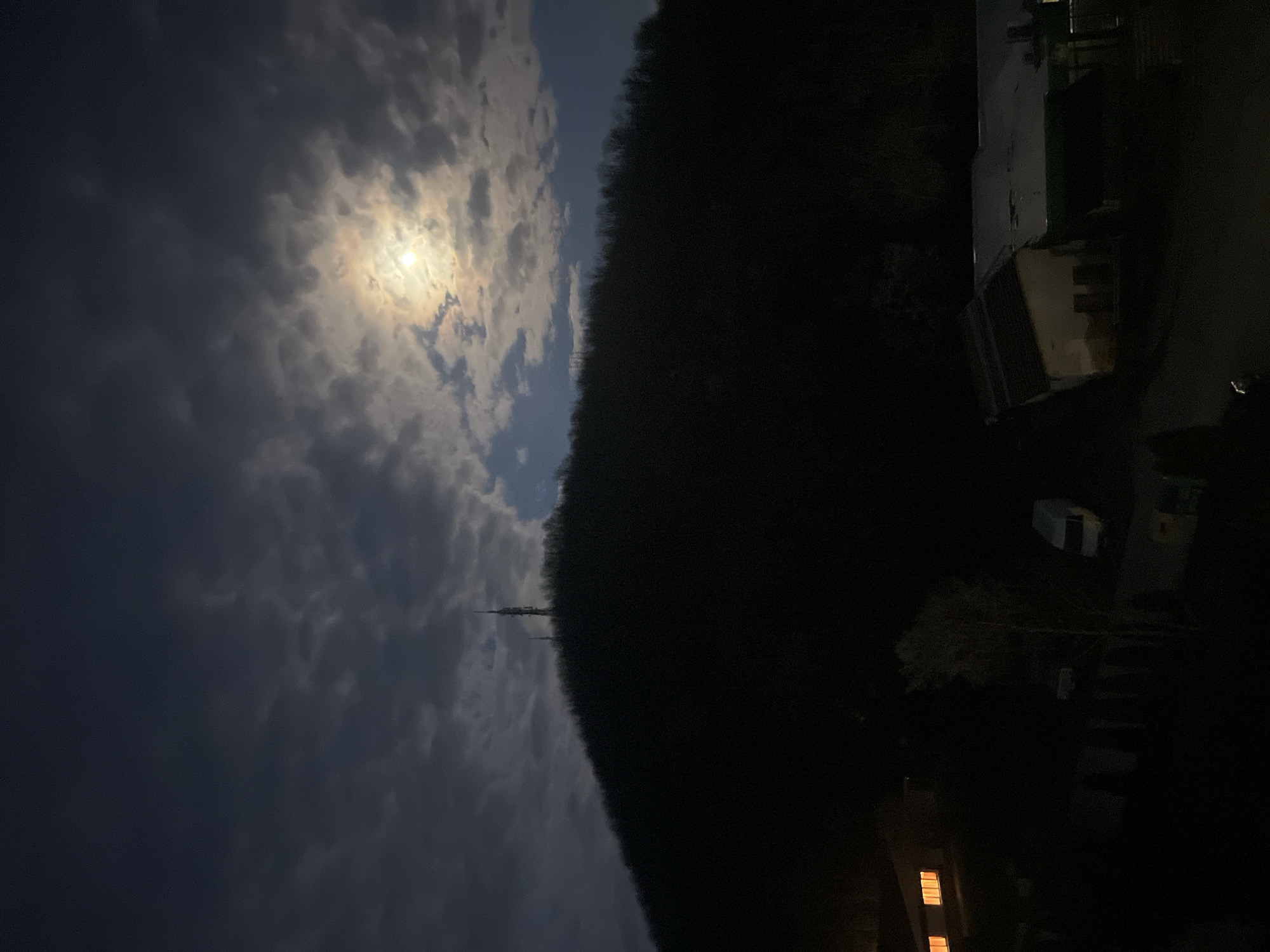 The image depicts a night scene in Elena, Bulgaria. The sky is partially cloudy, with the moon visible through a break in the clouds. A dark, forested hill dominates the right side of the frame. A transmission tower is silhouetted against the sky near the horizon. On the right side of the image, illuminated by artificial light, are buildings and a parked vehicle. The scene appears to be residential, with lights visible in the windows of a building in the foreground.