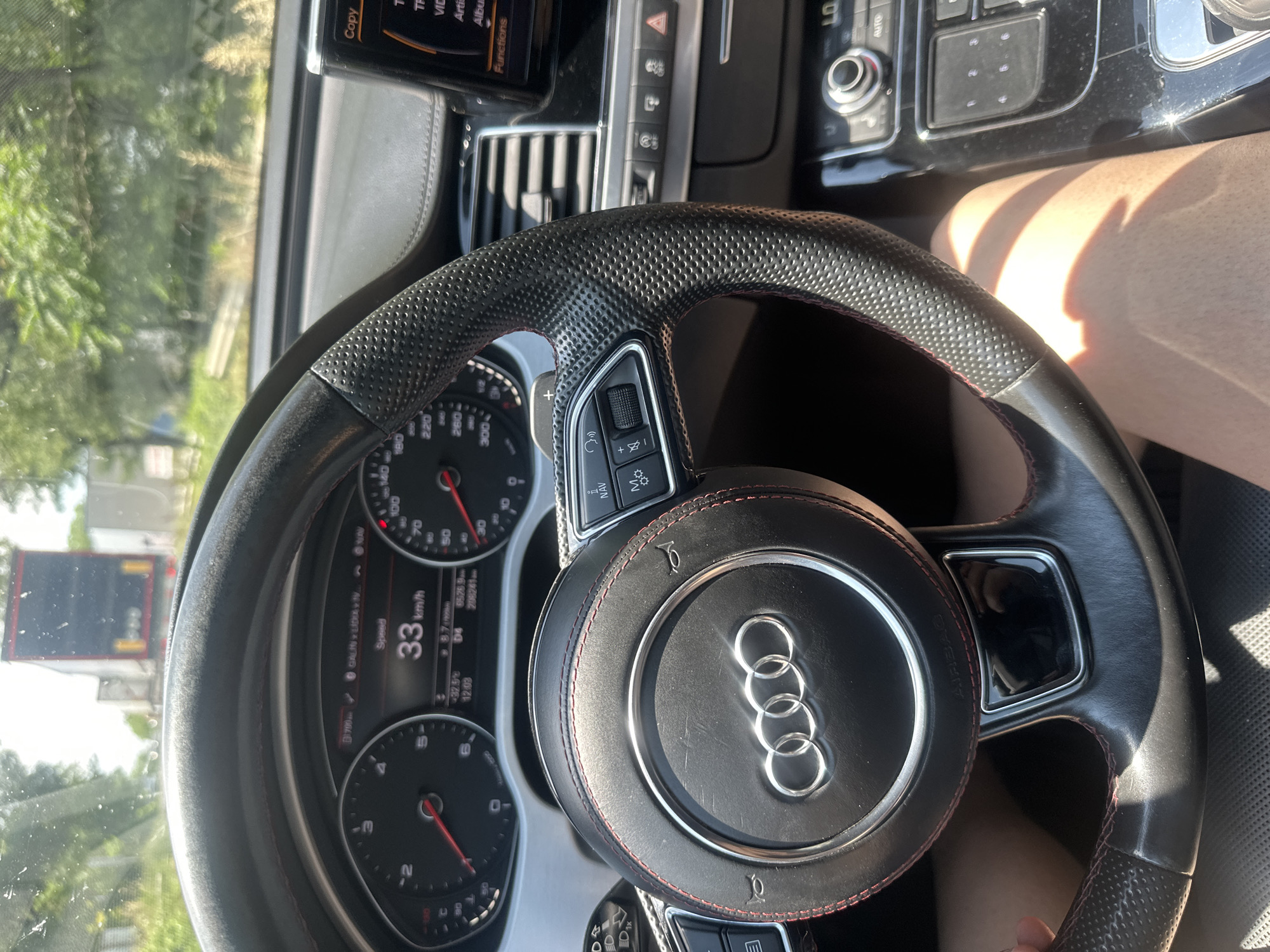 This media file shows a close-up view of the interior of an Audi car, focusing on the steering wheel and dashboard. The car appears to be in motion, as indicated by the speedometer displaying a speed of 33 km/h and the view of trees and a truck through the windshield. The driver's leg is partially visible on the right side of the frame, suggesting they are in the driver's seat. The dashboard features multiple displays, including a tachometer, speedometer, and a navigation screen showing "GALINLIDAN NAV". Sunlight is casting shadows on the dashboard and the driver's leg, indicating it is daytime. The steering wheel is black leather with red stitching and the distinctive Audi logo is prominently displayed in the center. Buttons and controls for the car's infotainment system are visible on the steering wheel and the center console. The scene appears to be set on a road in Polikraishte, Bulgaria.
---
The final answer is:
This media file shows a close-up view of the interior of an Audi car, focusing on the steering wheel and dashboard. The car appears to be in motion, as indicated by the speedometer displaying a speed of 33 km/h and the view of trees and a truck through the windshield. The driver's leg is partially visible on the right side of the frame, suggesting they are in the driver's seat. The dashboard features multiple displays, including a tachometer, speedometer, and a navigation screen showing "GALINLIDAN NAV". Sunlight is casting shadows on the dashboard and the driver's leg, indicating it is daytime. The steering wheel is black leather with red stitching and the distinctive Audi logo is prominently displayed in the center. Buttons and controls for the car's infotainment system are visible on the steering wheel and the center console. The scene appears to be set on a road in Polikraishte, Bulgaria.