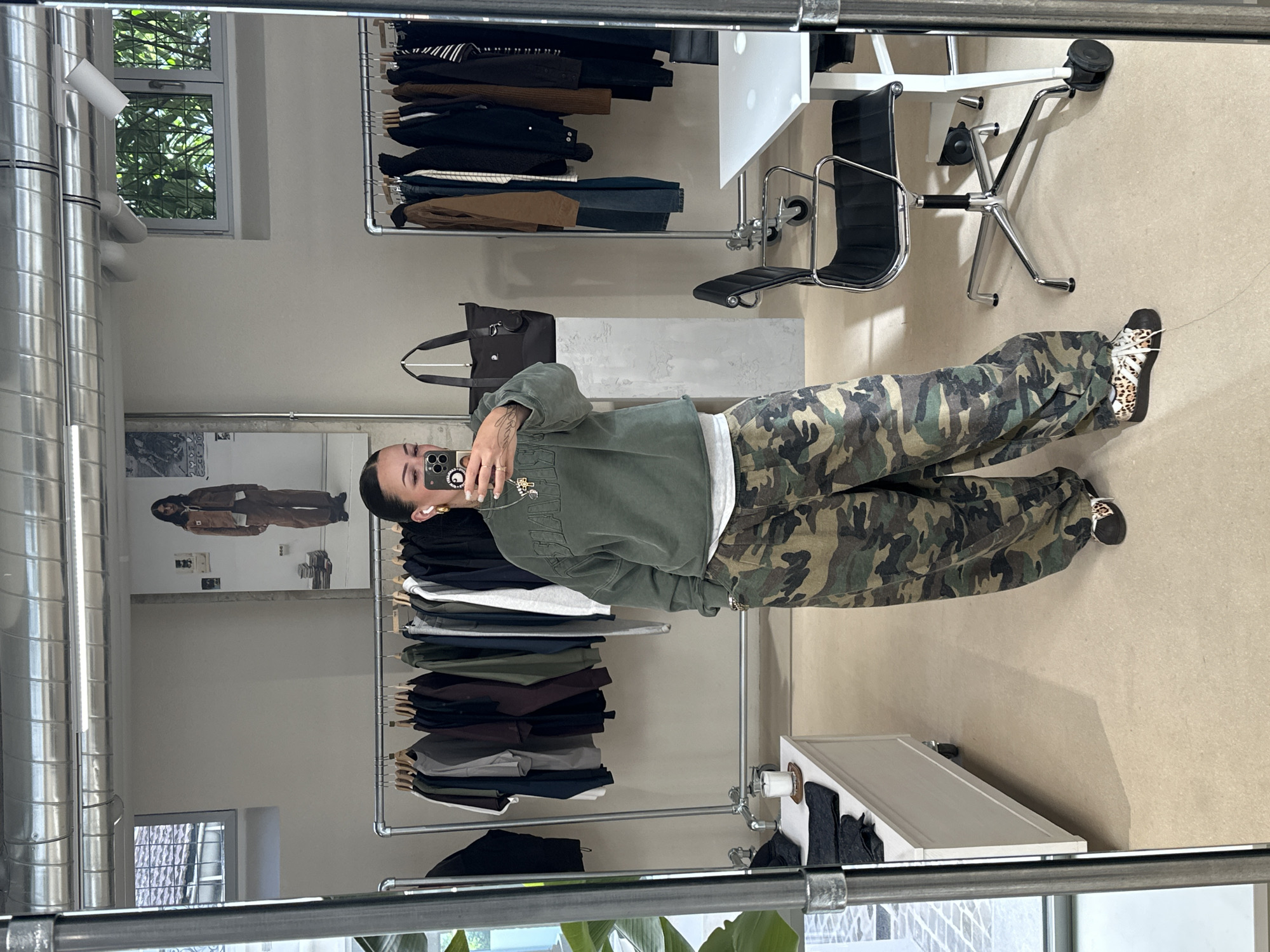 A reflection in a mirror shows a person wearing a green sweatshirt and camouflage pants. The person is holding a phone up to take the photo. The reflection includes clothing racks with various garments, a black leather bag, and office furniture including a desk and chair. The background features a window, interior wall, and other clothing. The setting appears to be a store interior with a modern aesthetic. There's no information to confirm the location as Paris, France.