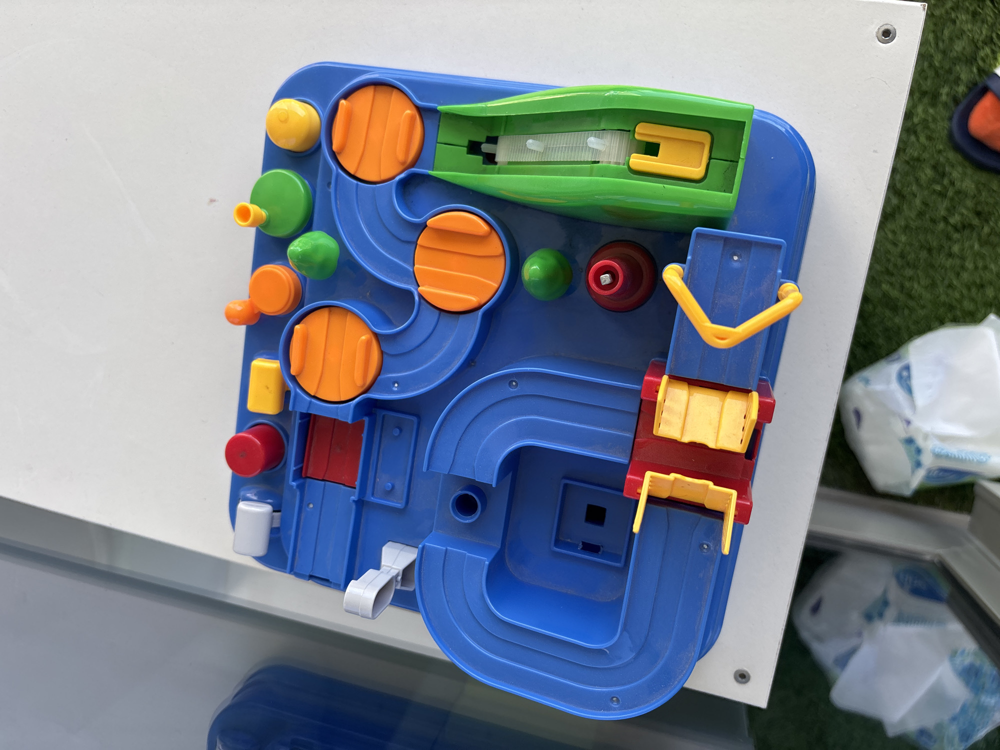The image shows a close-up, overhead view of a blue plastic children's toy with various colorful components, including orange ramps, green balls, yellow and red accessories, and a green track. The toy is positioned on a white surface, likely a table, which is placed on a green, artificial turf-like material. In the background, a reflection of a blue, curved object, possibly part of a larger structure or another toy, is visible. The overall setting appears to be indoors, with natural light illuminating the scene. There is no indication of the specific location within Dubai, United Arab Emirates.