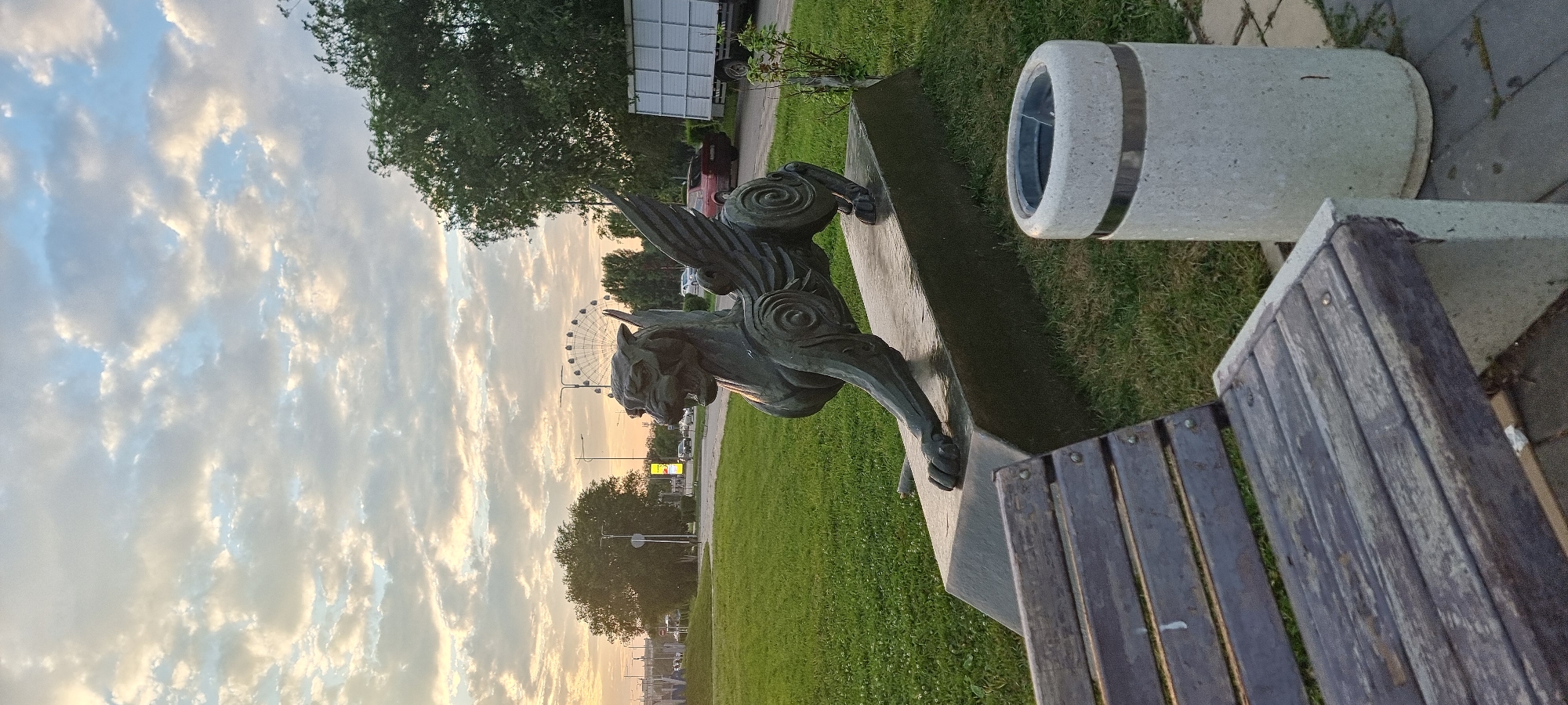 The image depicts a bronze statue of a mythological creature, possibly a griffin, situated on a concrete pedestal in a grassy park area. The statue is dark metallic in color and features wings, a fierce expression, and elaborate decorative swirls on its body and legs. In the foreground, a weathered wooden park bench is visible, and to the right, a cylindrical concrete trash bin with a metal band around its top. The background shows a street with moving vehicles, streetlights, and trees. Further in the distance, a Ferris wheel can be seen, suggesting the presence of an amusement park or fairground. The sky is filled with scattered clouds, illuminated by what appears to be the soft light of either sunrise or sunset. The location is identified as the area near the Aktobe Regional Mosque in the city of Aktobe, Kazakhstan.