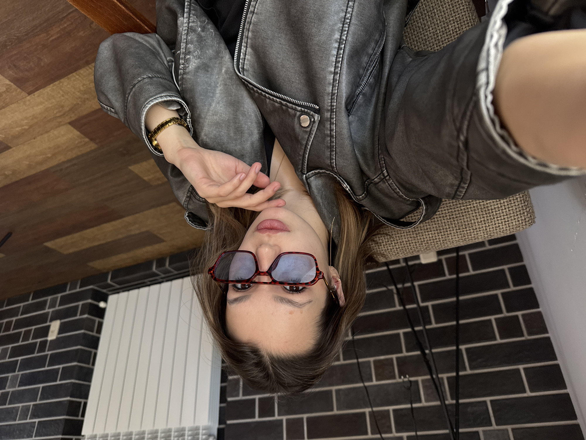 A young woman is shown in a selfie-style photograph. She is wearing rectangular sunglasses with a dark red frame. She also wears a distressed gray leather jacket. She has a gold bracelet on her left wrist and earrings.
The background includes a brick wall with dark gray bricks and lighter gray mortar. A white radiator is partially visible. Above, there is a wooden surface. Behind her is a textured neutral colored chair. There are some black cords running down the wall.