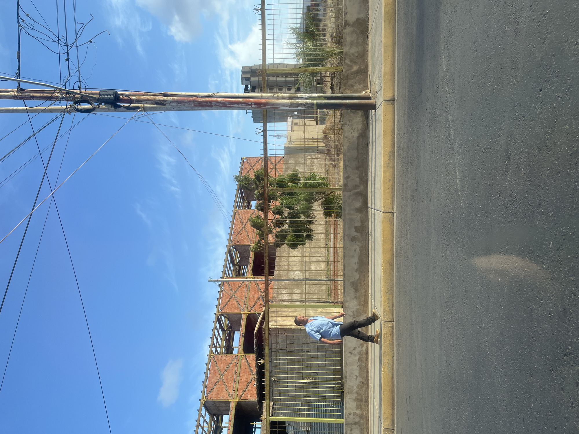 In Juan Griego, Venezuela, a man walks along a sidewalk in front of a construction site. The man is wearing a light blue collared shirt and dark pants, with brown shoes. He is walking from left to right across the frame. Behind him, a partially constructed brick building with exposed wooden framing is visible. A metal fence with a concrete base separates the sidewalk from the construction area. Further in the background, a taller, unfinished concrete building and some vegetation are present. The scene is illuminated by natural sunlight, casting shadows on the asphalt road in the foreground. A utility pole with numerous wires stands on the right side of the image, near the sidewalk. The sky is clear and blue with a few scattered clouds.