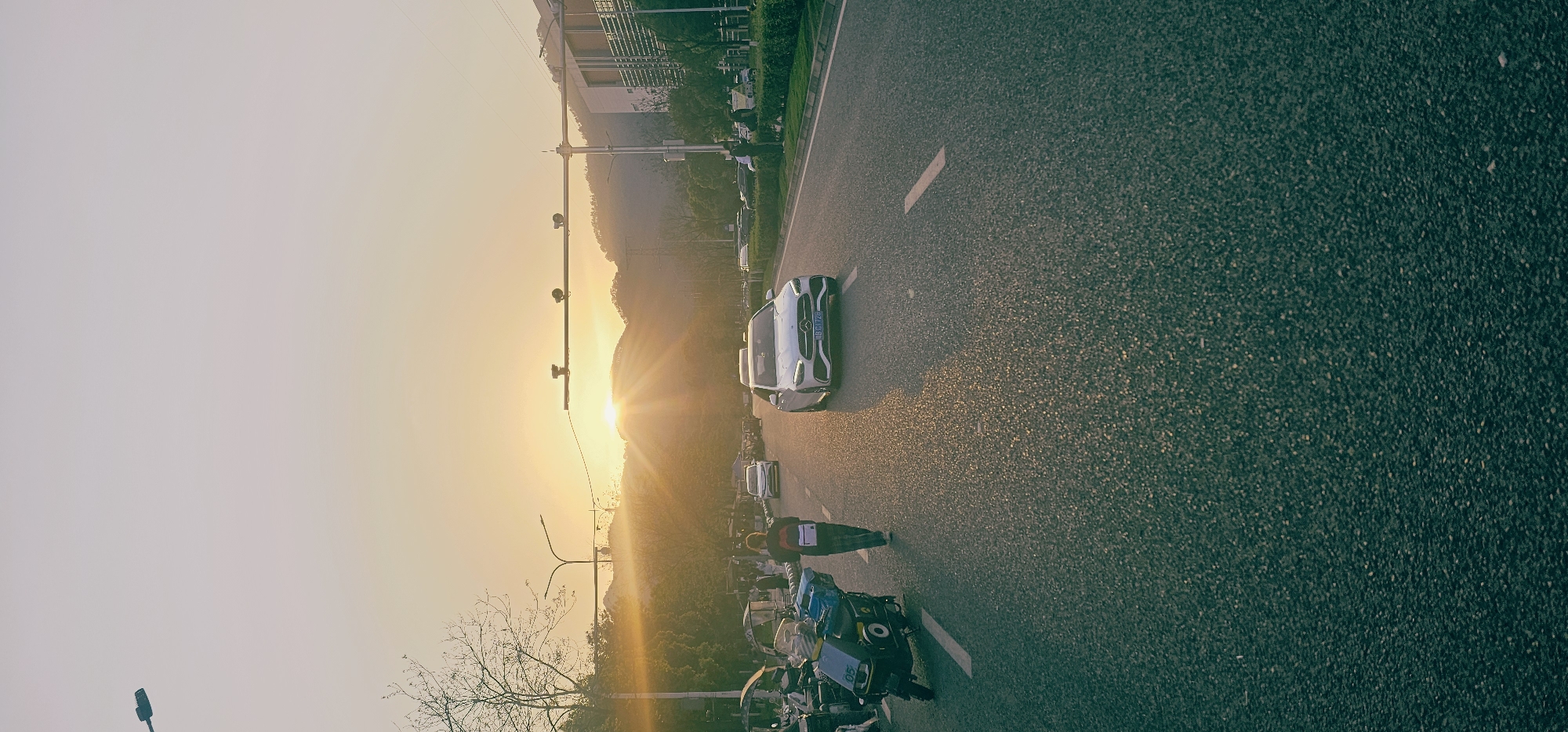 The image depicts a street scene at sunset. A silver Mercedes-Benz car is positioned on the right side of the road, which stretches from the bottom of the frame into the distance. A person is walking on the road near the car, away from the viewer. Two three-wheeled vehicles are situated on the roadside, close to the bottom of the image. Above the cars and people, the sun is setting behind a hill, with a light post and traffic lights in the foreground. The sky is bright orange and yellow near the horizon, fading into a lighter hue higher up. Buildings are visible on the left side of the road, along with lush trees. The location is Shapingba District, China, based on the license plate on the car.