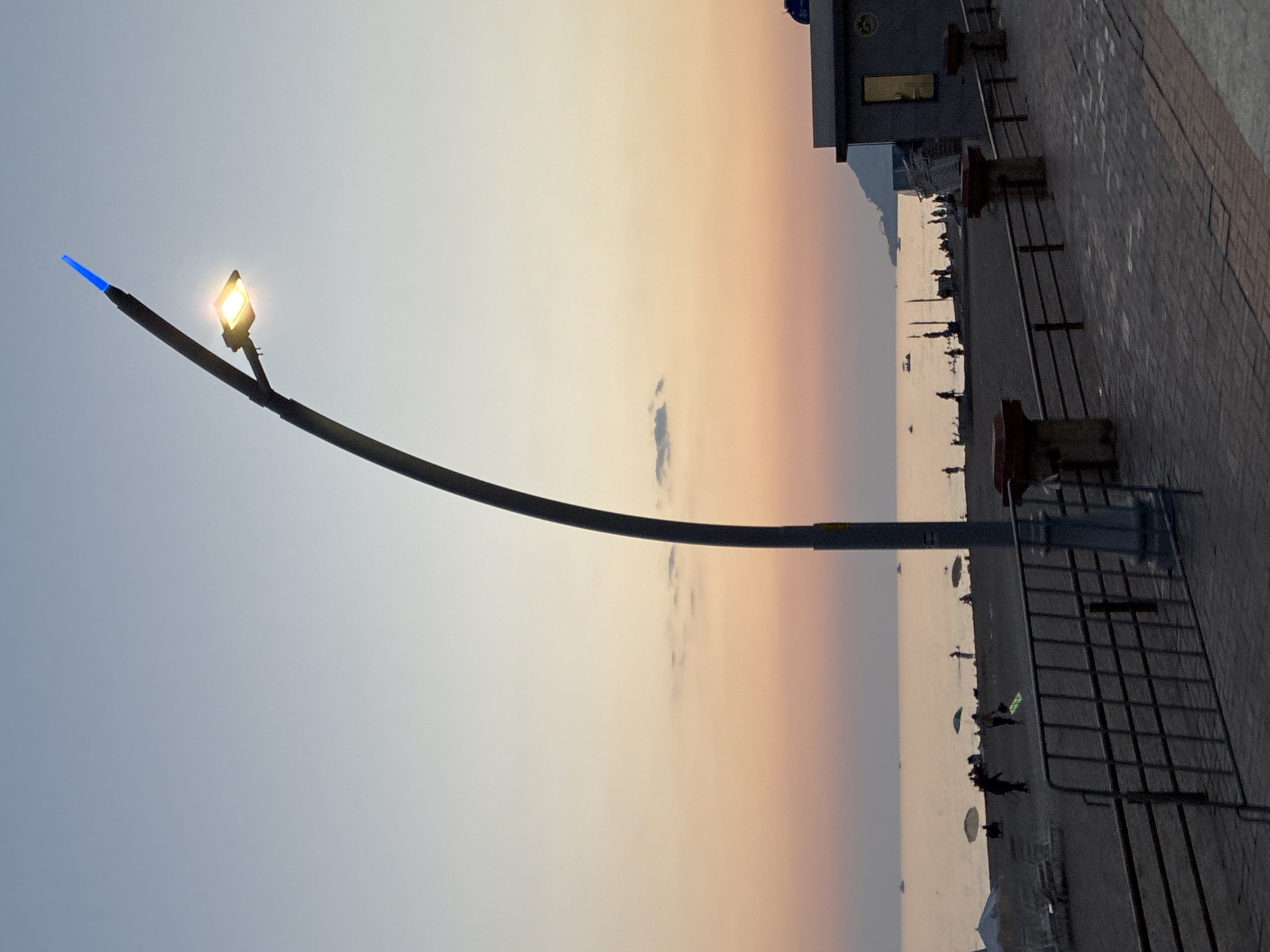 A tall, curved streetlamp with a lit yellow lamp and a blue tip stands prominently on a paved walkway overlooking a beach and the sea at sunset. The sky displays a gradient of soft blues and oranges, with a few wispy clouds. The beach is populated with people, some walking, some sitting, and some visible near beach umbrellas. The sea is calm with several small boats or buoys visible in the distance. To the right, a building with a window glows with an interior light, and a mountain is visible in the background. The foreground features a metal railing and the patterned brickwork of the walkway. The overall scene depicts a tranquil evening at a coastal location, likely in Alanya, Turkey, given the presence of a modern streetlamp design common in tourist areas.