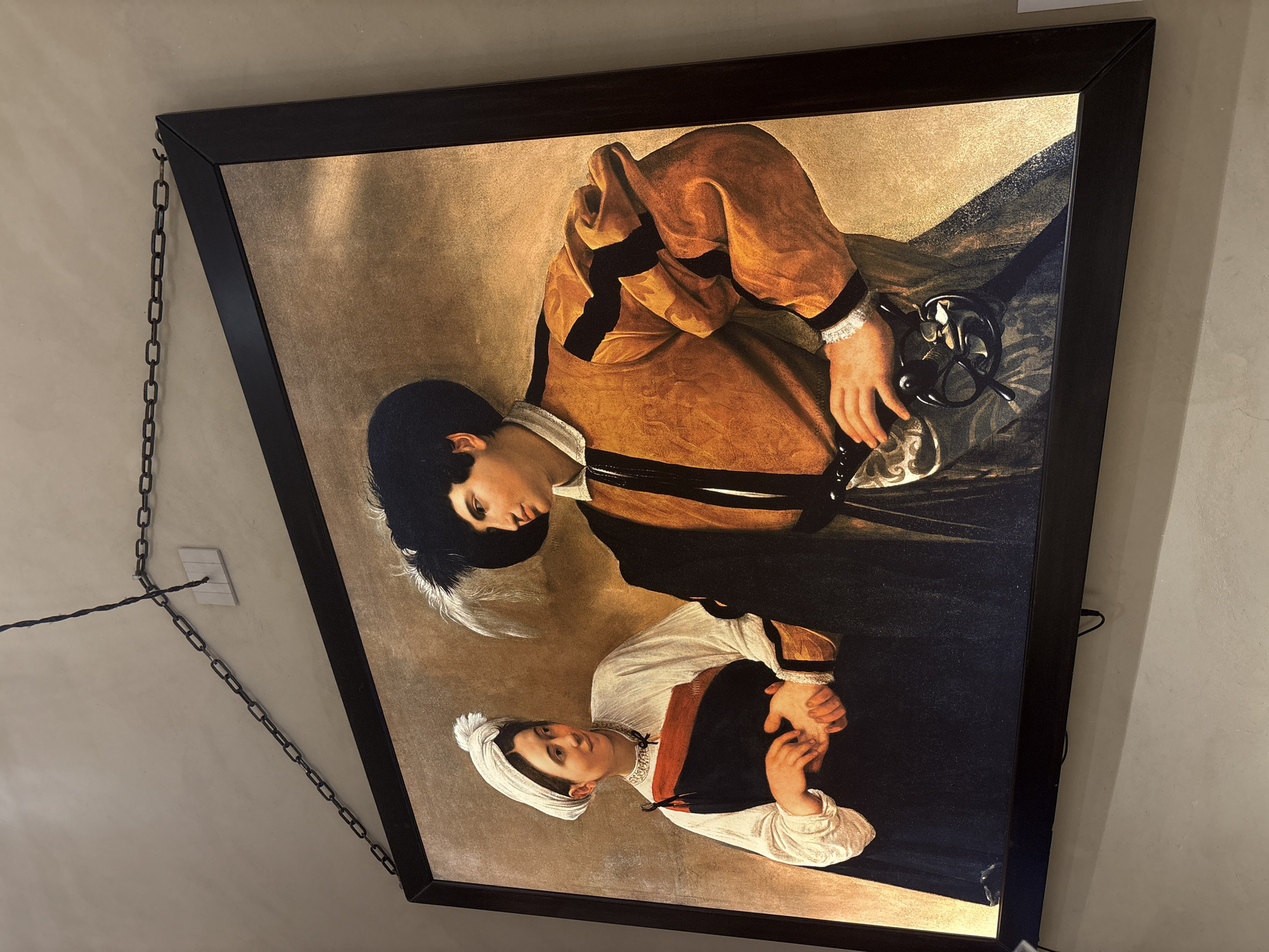 The image shows a framed painting of two figures, a man and a woman, indoors. The painting is hung on a light-colored wall by a dark metal chain. The man, on the right, wears a black hat with a feather, a yellow and black patterned jacket, and a white ruffled collar. He has one hand on his hip, which is adorned with a pouch and what appears to be a decorative sword hilt. The woman, on the left, wears a white head covering, a white long-sleeved shirt with a red and orange trim, and a dark blue garment. She is holding the man's hand with her left hand, and her right hand is resting on the man's hand. They are looking at each other, suggesting an interaction or connection. The background of the painting is a textured gold color. The painting is framed in a dark, simple frame. The room appears to be lit by artificial light, casting a warm glow on the painting. The location is not explicitly stated in the image, but it is implied to be in Rome, Italy.