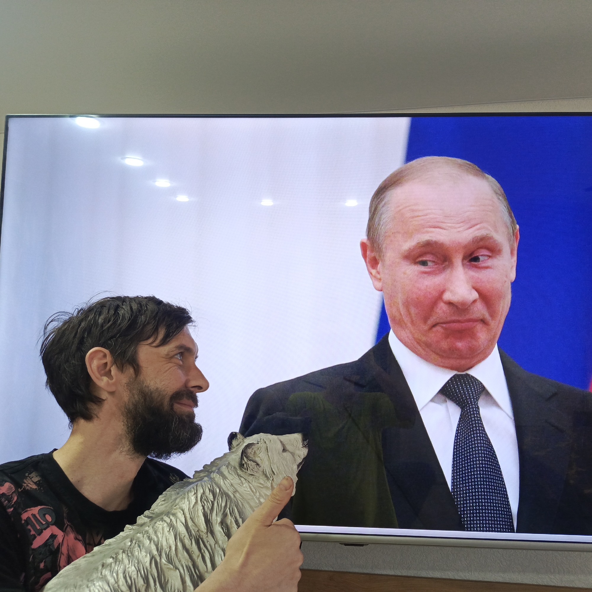 A man with a dark beard and hair stands to the left of a television displaying a video of Vladimir Putin. The man is holding a statue of a silver wolf, giving a thumbs up with his right hand near the screen. The video showing Putin features him in a dark suit, white shirt, and patterned tie. The background behind Putin is a blurred blue and white, possibly the Russian flag. The environment is indoors, appearing to be a casual setting. The image does not provide sufficient information to identify the location as Марковский, Russia.