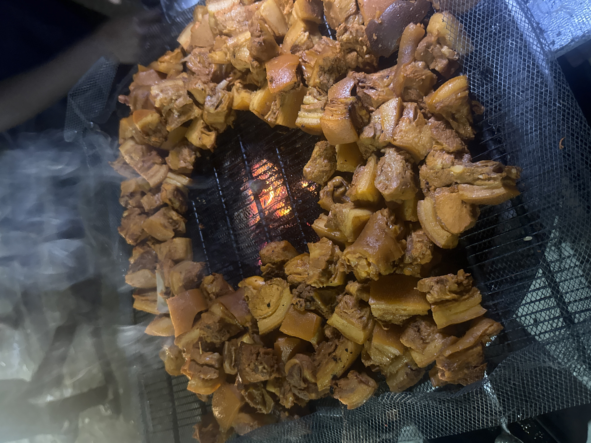 In Ihiagwa, Nigeria, a close-up shot captures a collection of roasted pork pieces arranged in a circular pattern on a metal grill. The pork, cut into irregular chunks, exhibits a rich brown color with visible layers of fat and meat, indicating it has been cooked over a heat source, likely charcoal or wood, as evidenced by the glowing embers visible beneath the grill. Wisps of steam rise from the hot meat, suggesting it is freshly cooked or still warming. The grill itself is placed on a mesh surface, possibly a makeshift cooking setup. In the background, blurred figures and smoky conditions suggest an outdoor or open-air cooking environment, possibly a street food stall or a local gathering. The lighting is dim, focusing attention on the textured details of the pork.