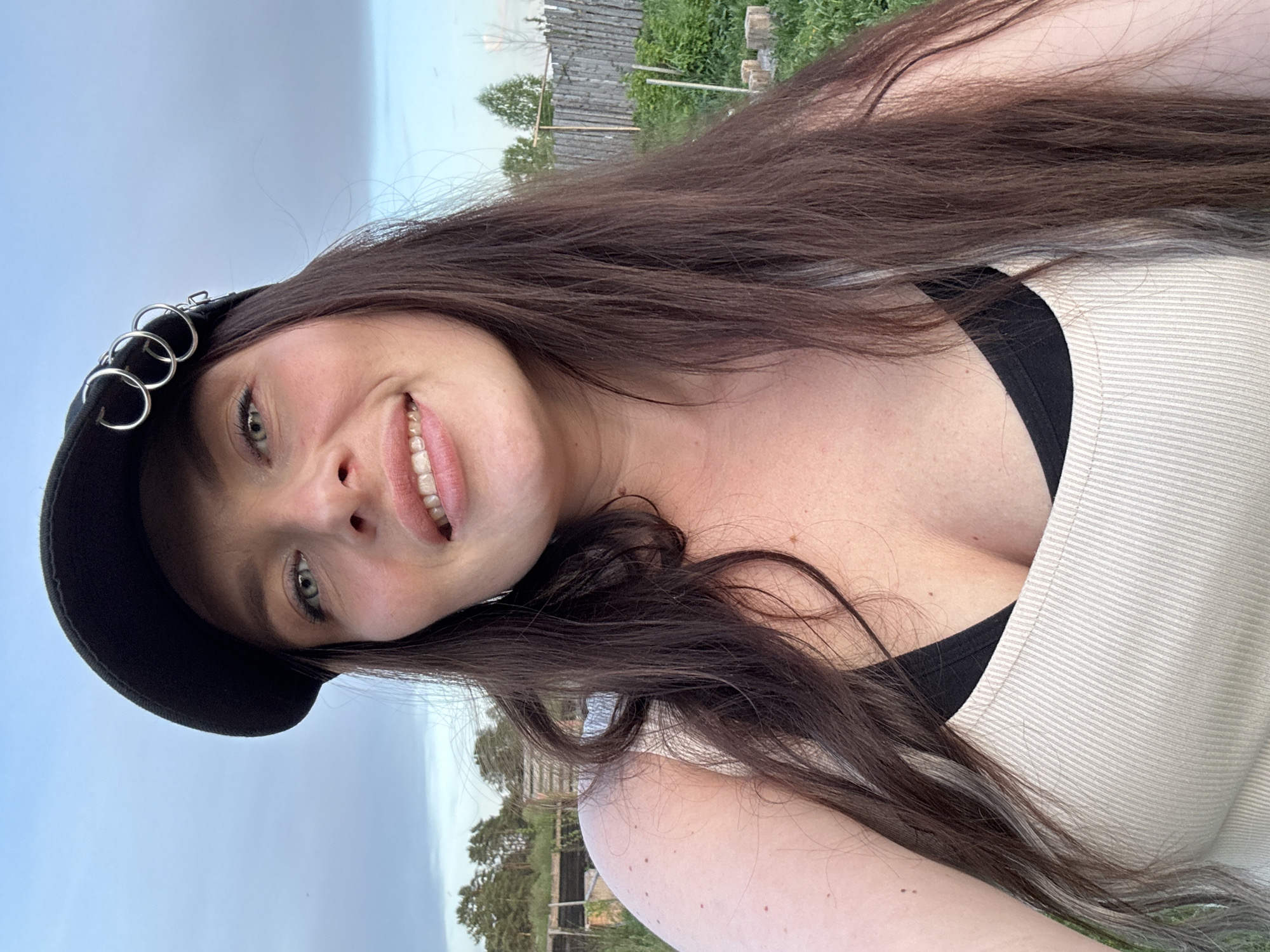 A woman with long, dark brown hair smiles while wearing a black hat with silver rings. She wears a light-colored tank top over a black undergarment. The background shows a grassy area and a wooden fence under a cloudy sky. The location is likely outdoors based on the vegetation and sky. There is no specific information to confirm the location is Rusalevka, Russia.