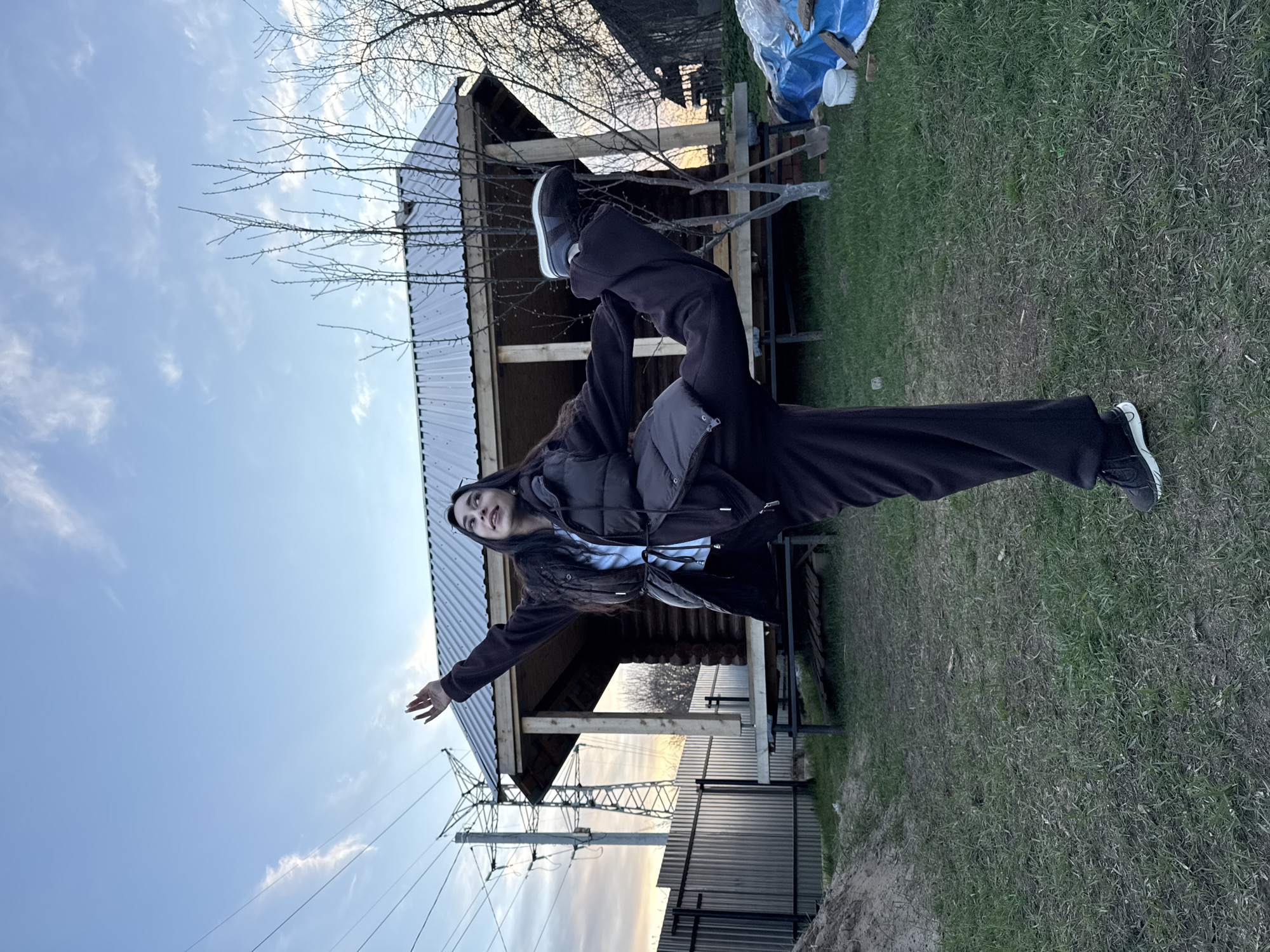 The image captures a young individual striking an athletic pose outdoors in Chaikovsky, Russia, during what appears to be dusk or dawn. The subject, a person with long dark hair, wears a dark puffer vest over a dark long-sleeved top, dark wide-leg pants, and dark athletic shoes with white soles. They are balanced on one leg, with the other lifted high and bent at the knee, while their arms are extended outwards for balance. A soft smile is visible on their face as they look slightly upwards.
In the background, a wooden structure, possibly a house or shed under construction, features a corrugated metal roof. Bare tree branches are visible against the sky. The ground is covered with green grass, appearing somewhat sparse in areas. To the right, a blue tarp covers items, and a white bucket is present. Further in the distance on the left, an electricity pylon with power lines stands against the horizon. The sky is a clear blue, transitioning to lighter tones near the horizon with scattered white clouds, indicating the early morning or late evening light. There is no visible text in the image.