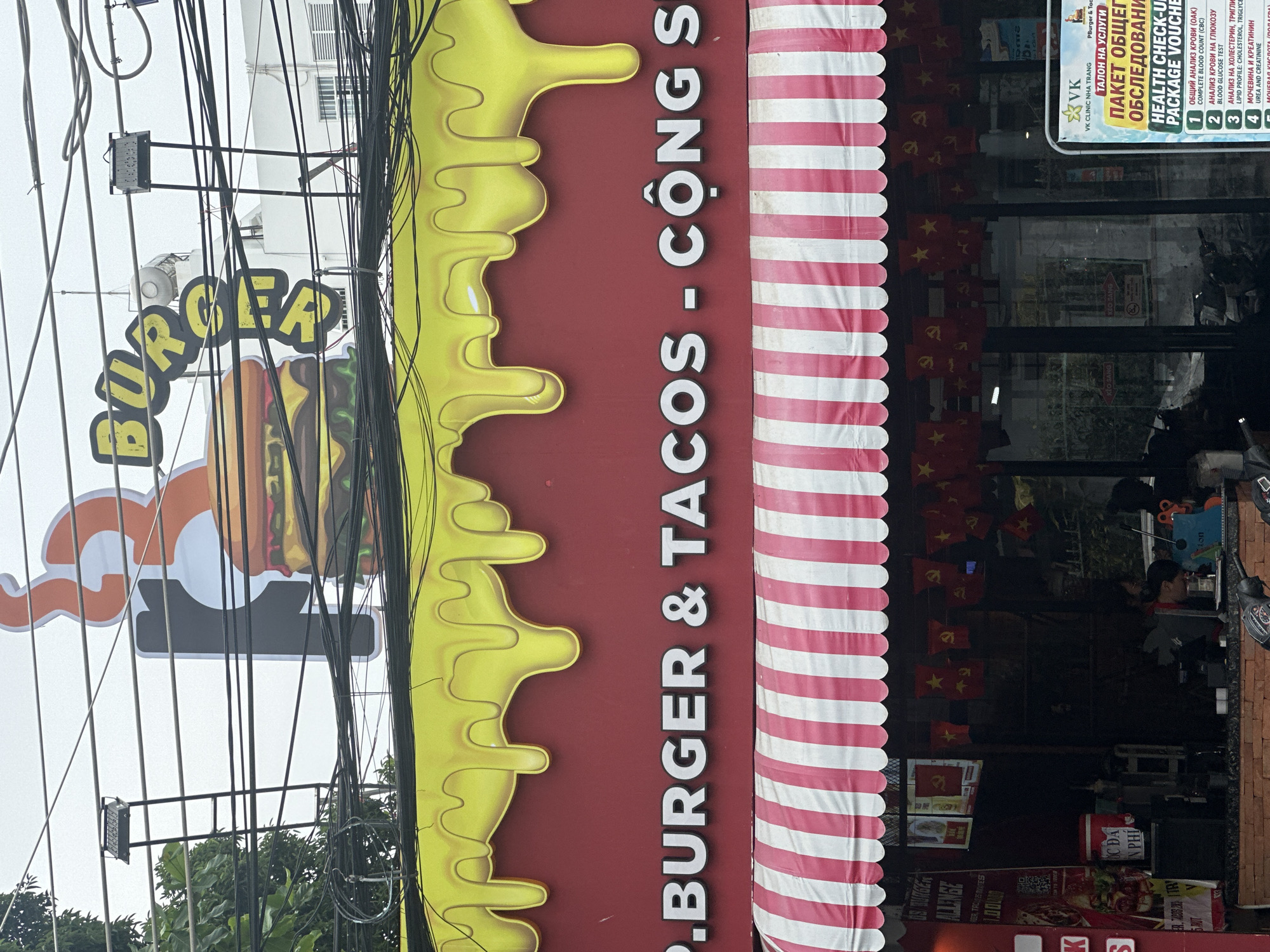 Here is a description of the image:
The image depicts a street scene in Bac Nha Trang Ward, Vietnam, with a focus on a restaurant called "Burger & Tacos - Cộng S". The restaurant's sign is prominently displayed, featuring the restaurant's name in white letters against a maroon background, with a stylized yellow drip effect on the left and a red and white striped pattern on the right. A smaller burger sign can be seen with black wires over it. The entrance to the restaurant is visible, decorated with Vietnamese flags, and there are people inside. There is also another sign in front advertising "Health Check-Up Package Voucher" from a clinic. The surrounding environment includes power lines and lush greenery, which are commonplace in this area.