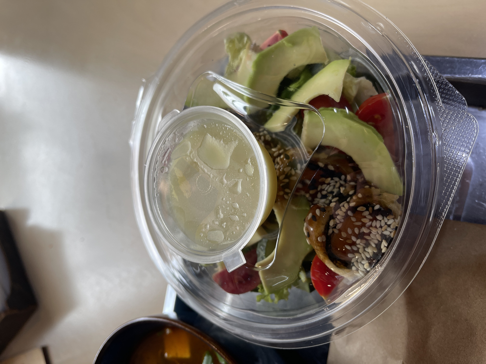 A clear plastic container holds a salad with avocado slices, cherry tomatoes, and what appears to be sushi rolls drizzled with a dark sauce and sprinkled with sesame seeds. A small, clear plastic cup with a light-colored dressing is placed on top of the salad. To the left of the salad container is a dark bowl containing a broth-based soup with some green garnish. The objects are situated on a light-colored, textured surface, possibly a table.