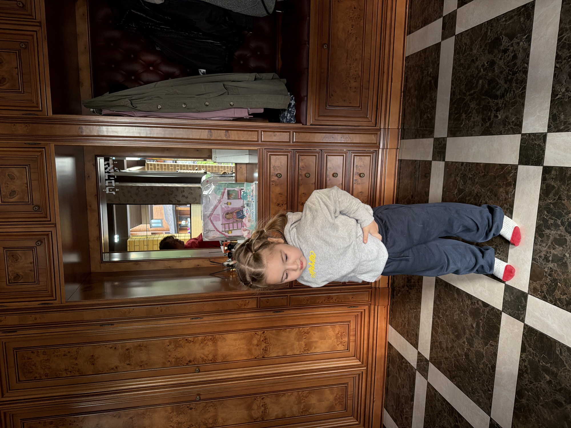 A young child stands in front of a large, wooden wardrobe in a room with a black and white checkered floor. The wardrobe has multiple sections including drawers, cabinets, and a space at the top where clothes are hanging. A mirror is affixed to the wardrobe. The child is wearing a gray sweatshirt and dark blue pants with red and white socks. The mirror is reflecting a room, presumably the outside. The location is inferred to be Новоглаголево, Russia.