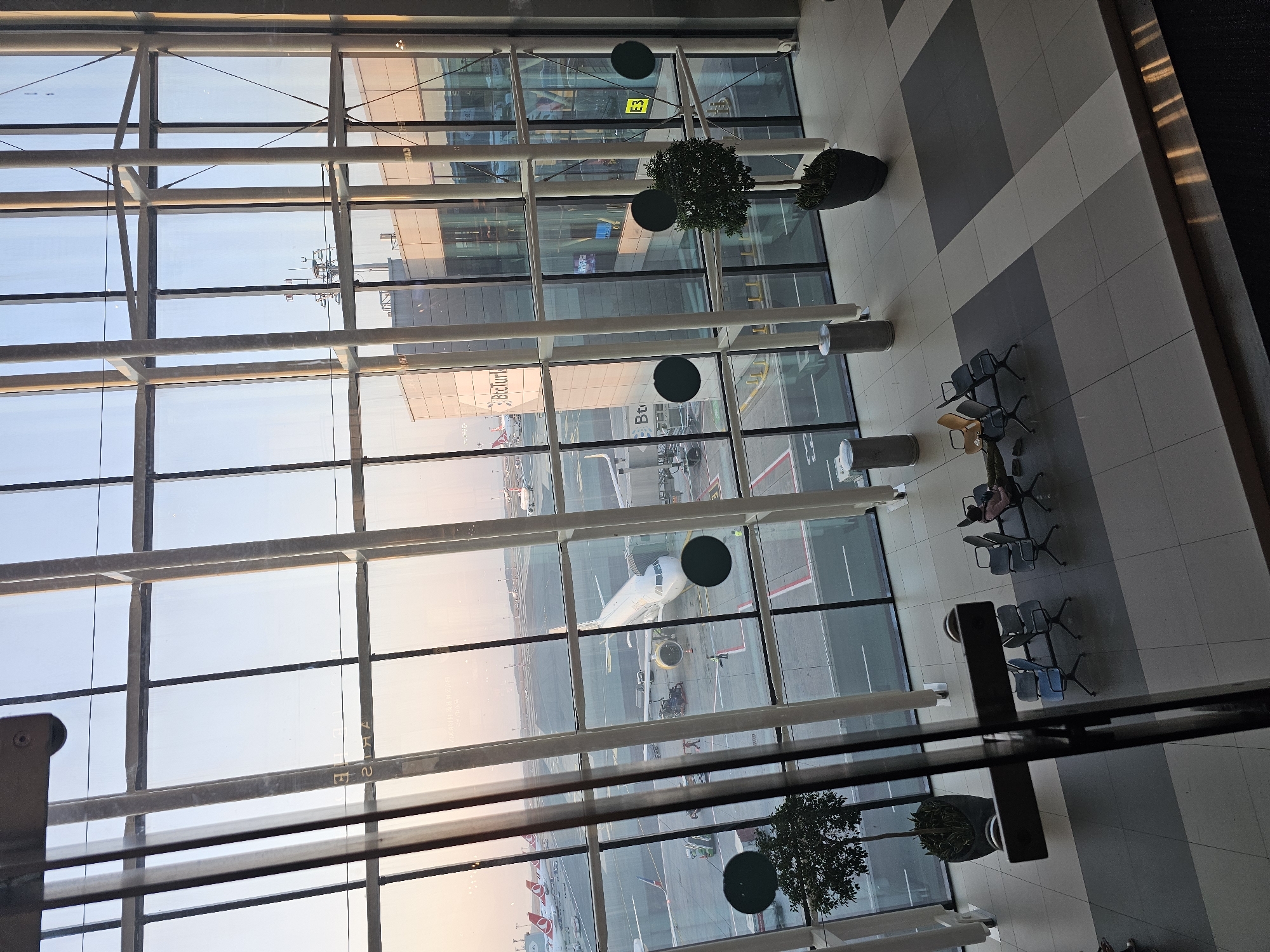 The image captures an interior view of an airport terminal in Arnavutköy, Turkey, looking out onto the tarmac through a large glass-paneled wall. The scene is bathed in the warm glow of what appears to be sunrise or sunset, casting long shadows and highlighting the architectural elements.
Inside the terminal, several rows of empty, modern chairs are visible on a tiled floor, with some areas featuring darker and lighter grey tiles. Two potted trees, one on the left and one on the right, add a touch of greenery to the space. Two people are seated in the chairs on the right side of the image, one with their head down, seemingly resting or sleeping. Two cylindrical trash receptacles are also present.
Outside the terminal, a white airplane is parked at a gate, with a jet bridge connected. Ground crew members are visible working around the aircraft. Further in the distance, other airplanes are parked on the tarmac, and the runway stretches out towards the horizon. The sky beyond is a pale blue with hints of pink and orange near the horizon. The architectural style of the terminal appears modern and functional, with prominent vertical and horizontal structural elements supporting the glass facade. A sign with the text "BTCTURK" is visible on the exterior of the terminal building. The overall impression is one of calm and order within a busy transportation hub.