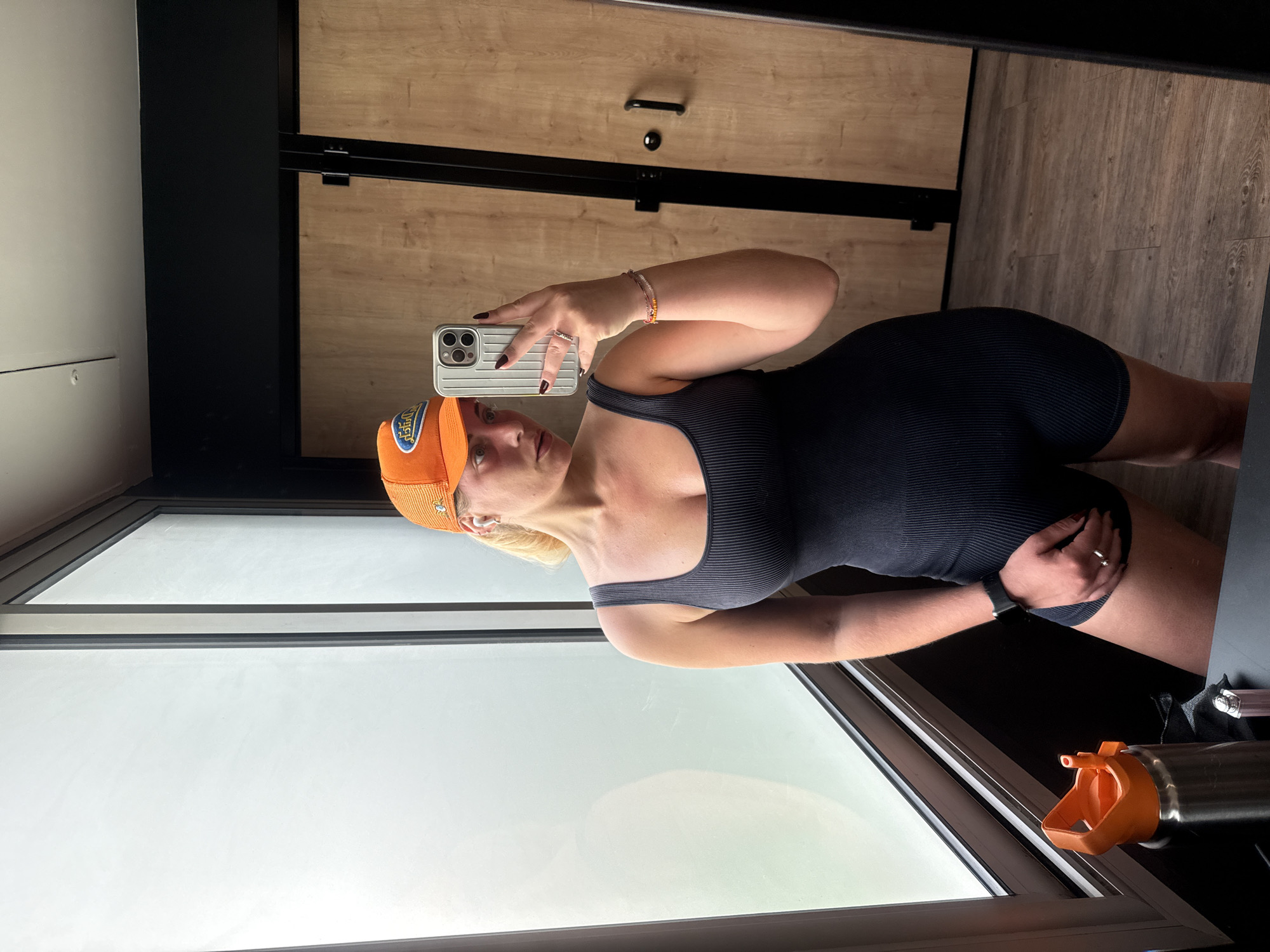 A woman is posing for a selfie in front of a window. She is wearing a dark gray ribbed tank top and matching shorts. She is also wearing an orange baseball cap. The woman is holding a cell phone in her right hand. Her left hand is resting on her leg. A water bottle is on the window sill. The background consists of a light-colored cabinet and a black frame. There is no indication of the location being Paris, France.