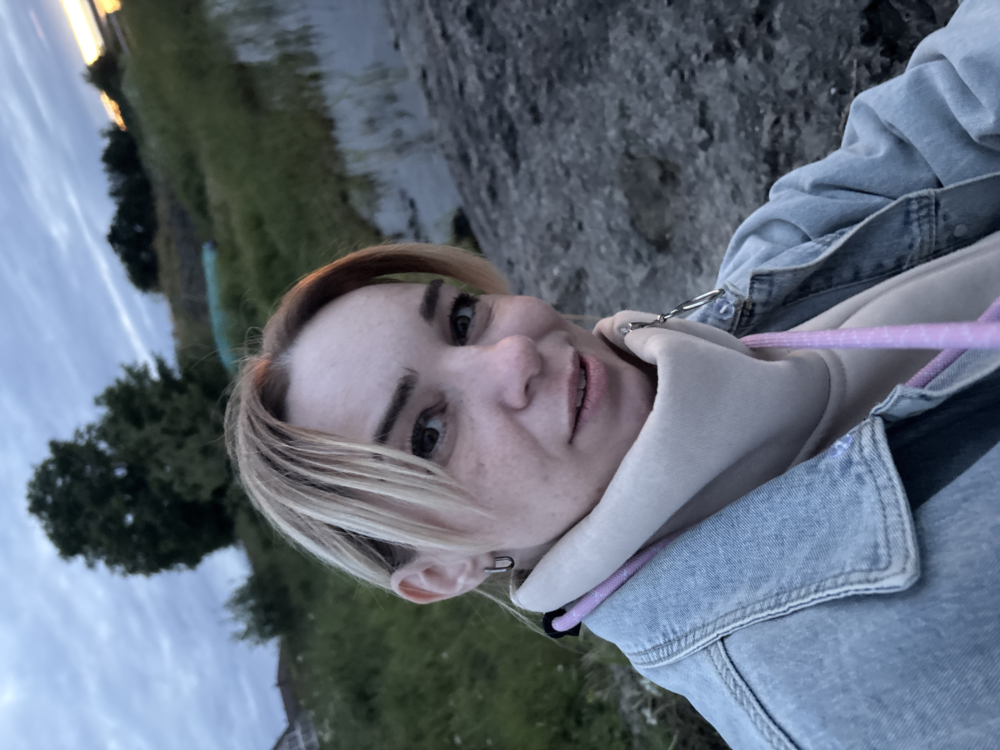 A medium-shot, natural-light, outdoor selfie shows a young Caucasian woman with blonde hair and freckles looking directly at the camera with a slight smile. She is wearing a light blue denim jacket over a cream-colored hoodie. A pink drawstring is visible around her neck. She has a silver hoop earring in her left ear. The background features a body of water on the right, with a rocky shore in the foreground. Beyond the water, there is a grassy bank with trees and a building on the left, under a cloudy sky with hints of sunset on the horizon. The setting appears to be a natural landscape near a body of water, possibly a lake or river, on the outskirts of Shuiskaya Chupa, Russia.